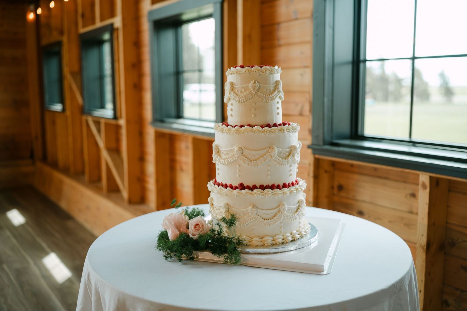 Elegant three-tier white wedding cake with red berry accents and floral decorations on a round table in a rustic barn venue.
