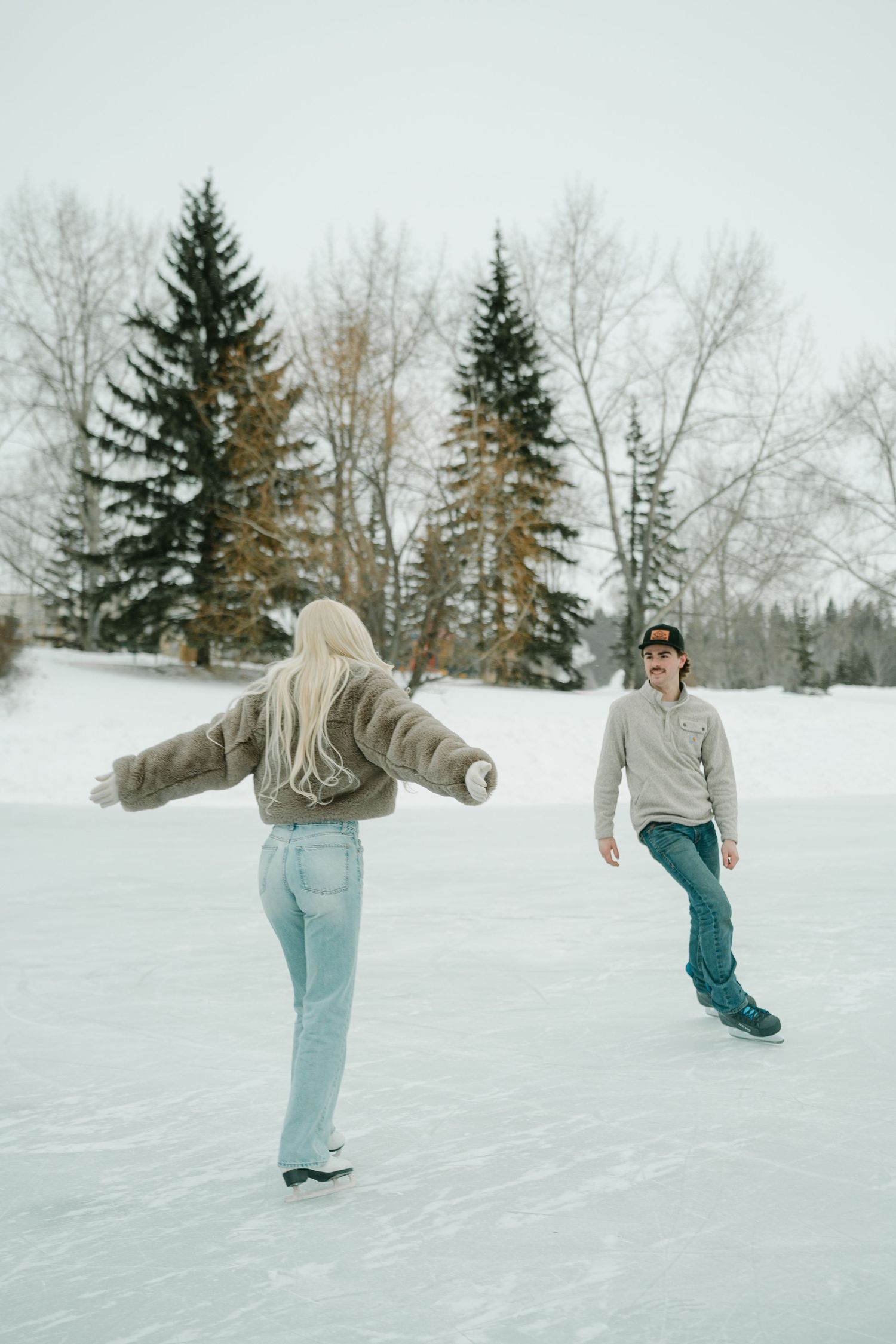 Couple ice skating outdoors in winter, surrounded by snow-covered trees. Bower ponds Red Deer Alberta. Engagement photos.