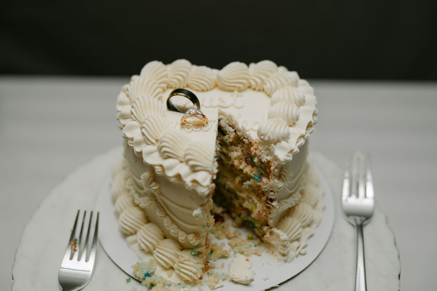Heart-shaped wedding cake with white frosting and gold rings on top, with a slice removed revealing colorful interior.