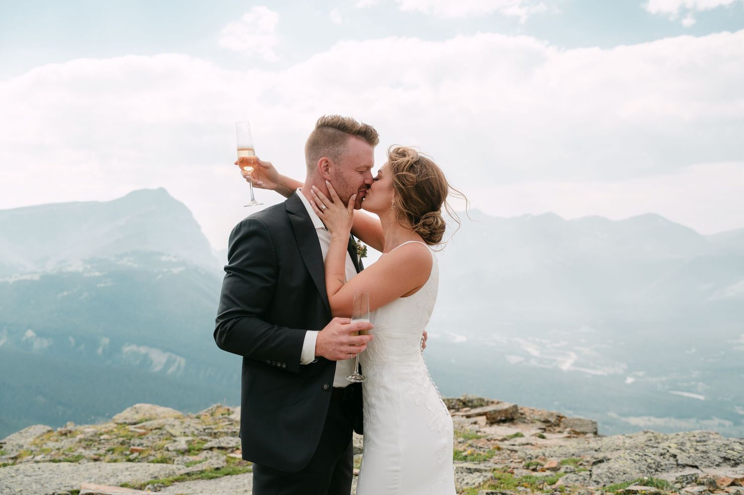 Bride and groom kissing on mountain summit holding champagne with scenic Rocky Mountain backdrop.