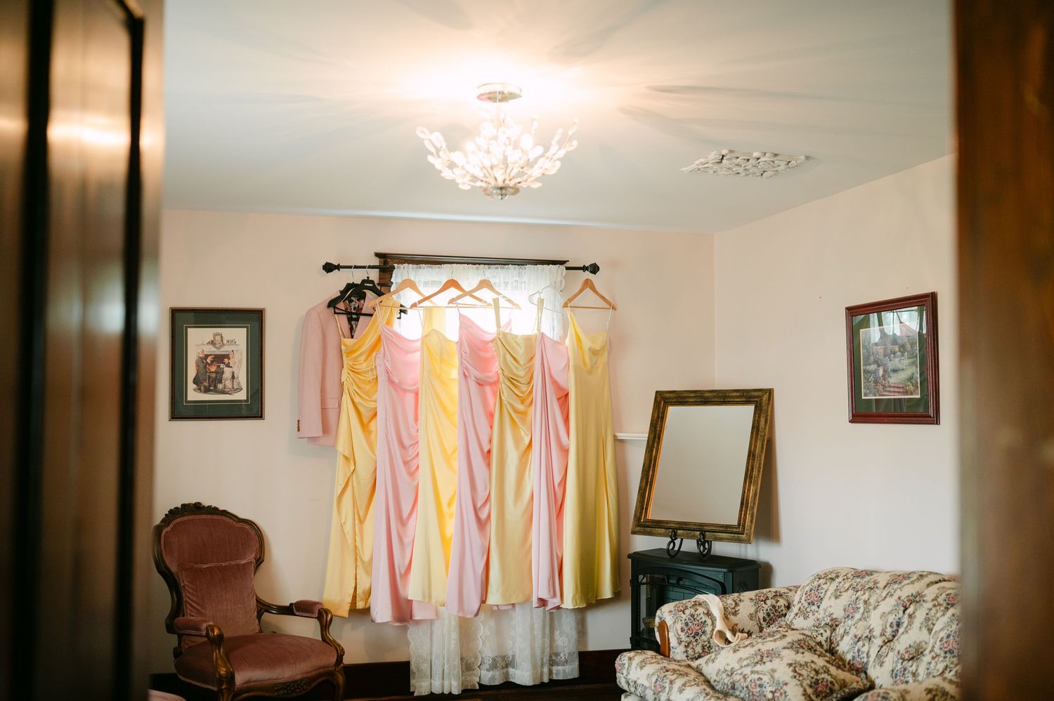 Pink and yellow bridesmaid dresses hanging on a rod in an elegant room with a chandelier and vintage furniture.