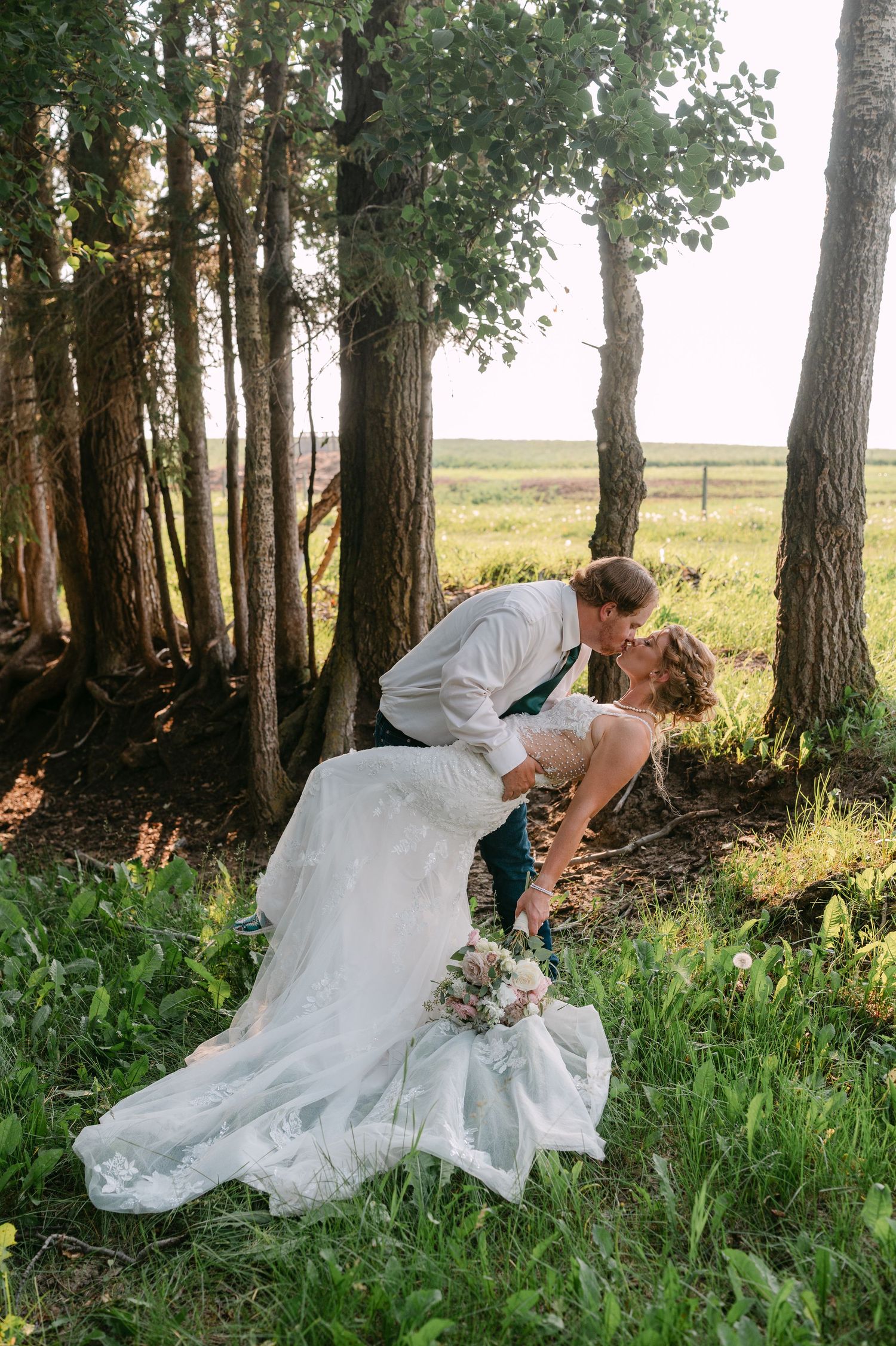 Bride and groom share a romantic dip kiss among tall trees in a lush green field during outdoor wedding photos.