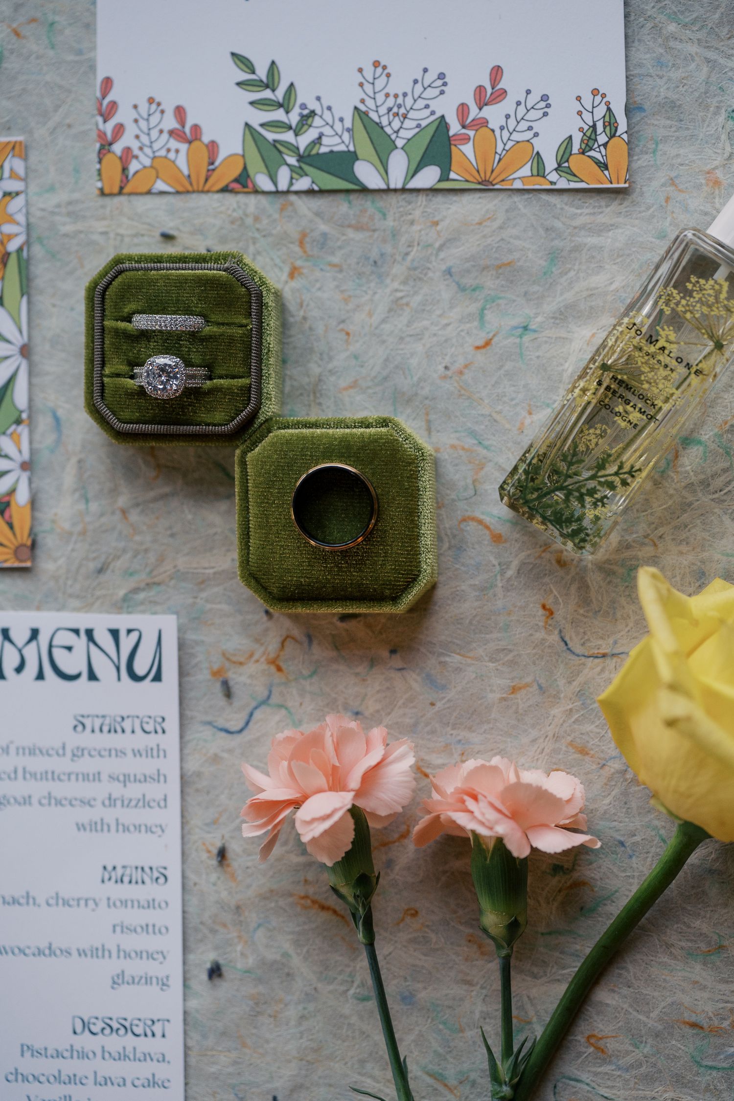 Wedding flat lay featuring diamond rings in green velvet box, floral menu, and pink carnations.
