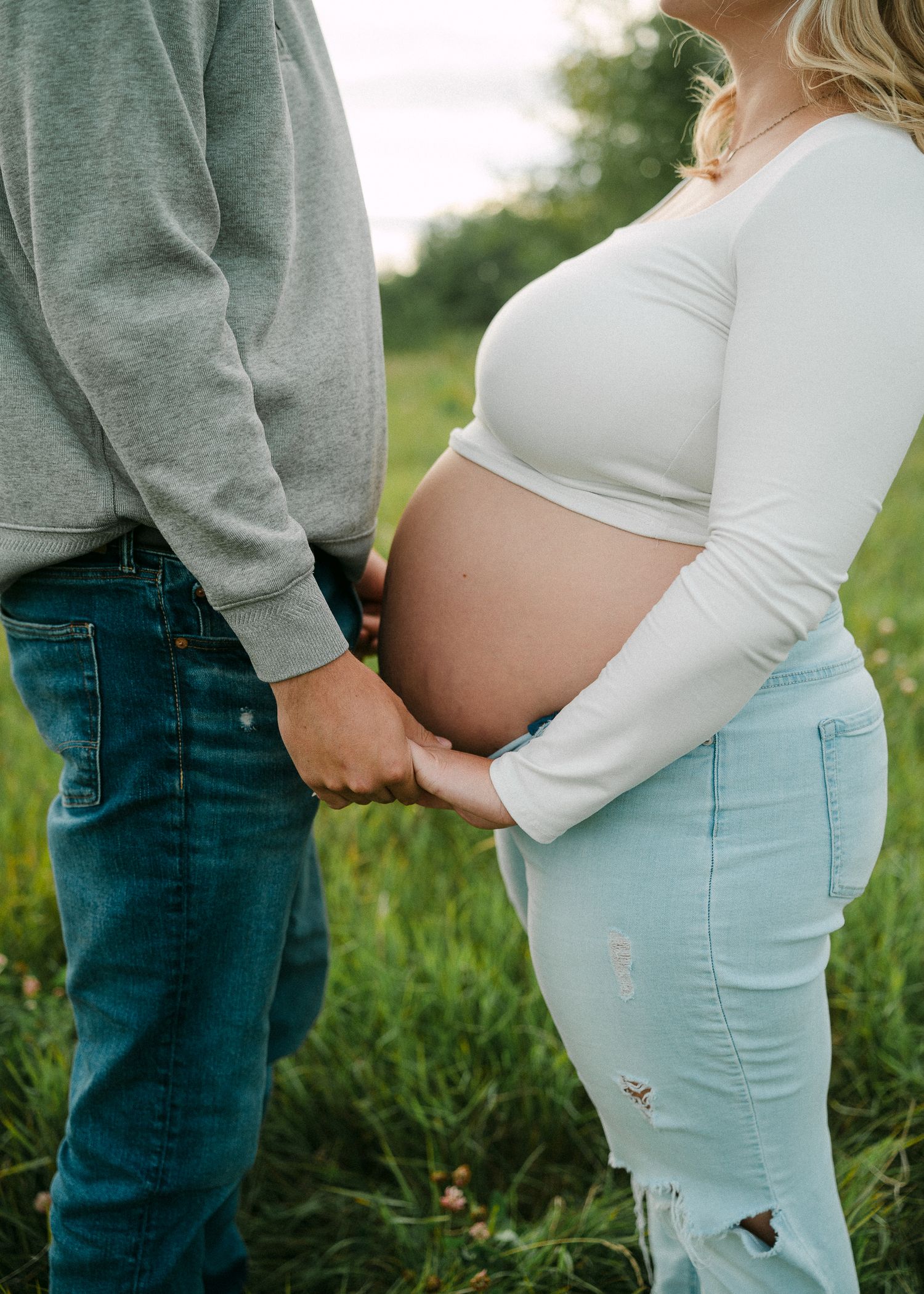 Pregnant woman in white crop top and light jeans holds hands with partner in green field during maternity photoshoot.