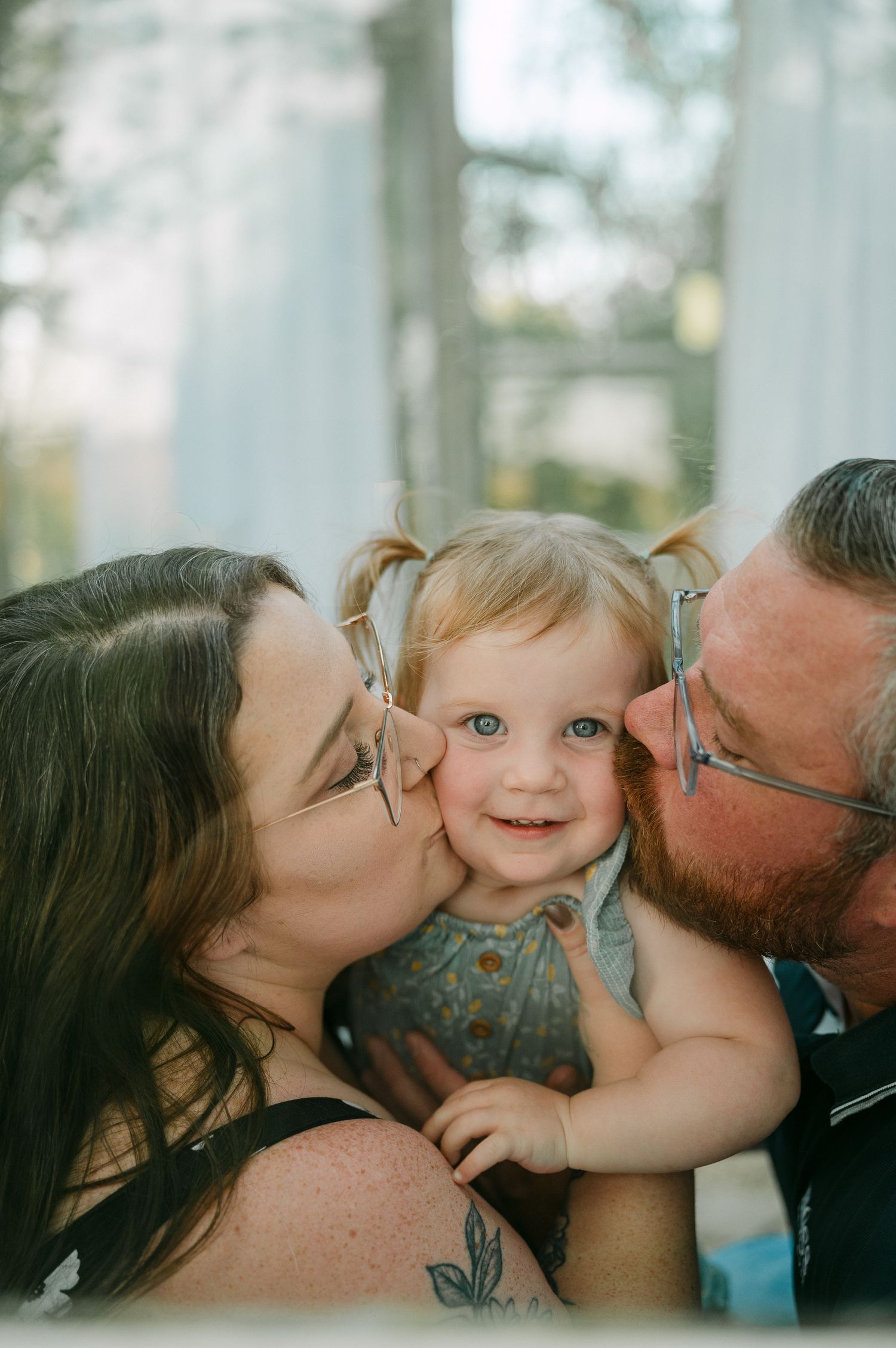 Two parents with glasses kissing the cheeks of a smiling baby girl with pigtails. family mini session