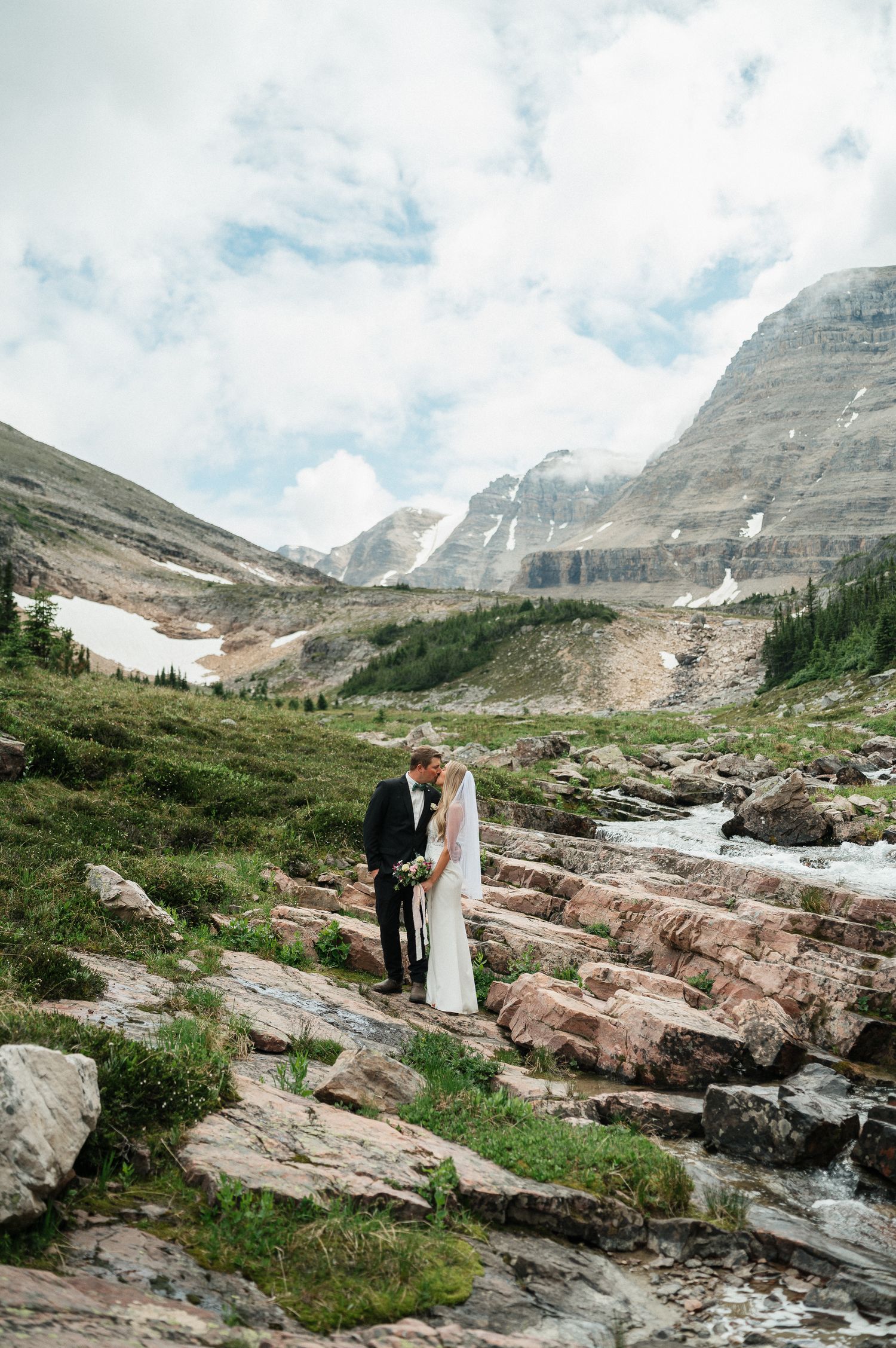 Bride and groom share a romantic moment on rocky terrain with stunning mountain backdrop and stream.