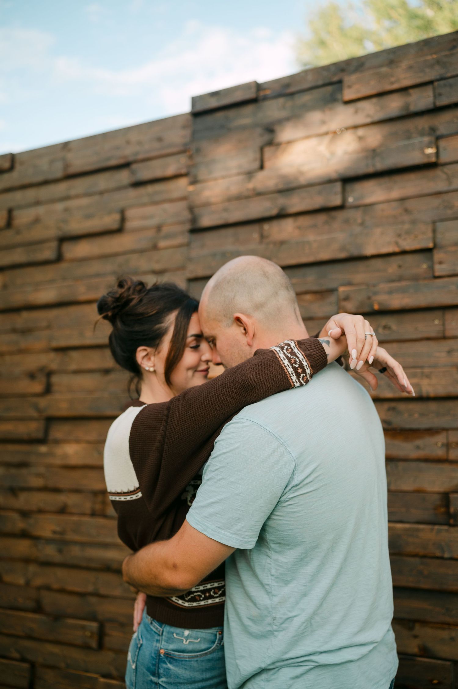 Couple embracing tenderly in front of a rustic wooden wall outdoors, sharing an intimate moment.