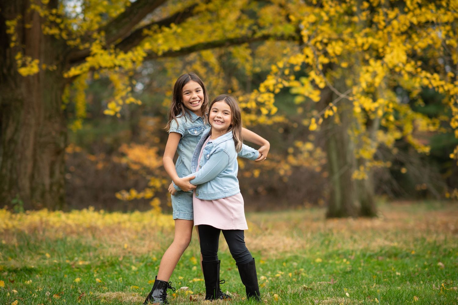 Two girls hugging and smiling in a park with vibrant yellow autumn foliage in the background.