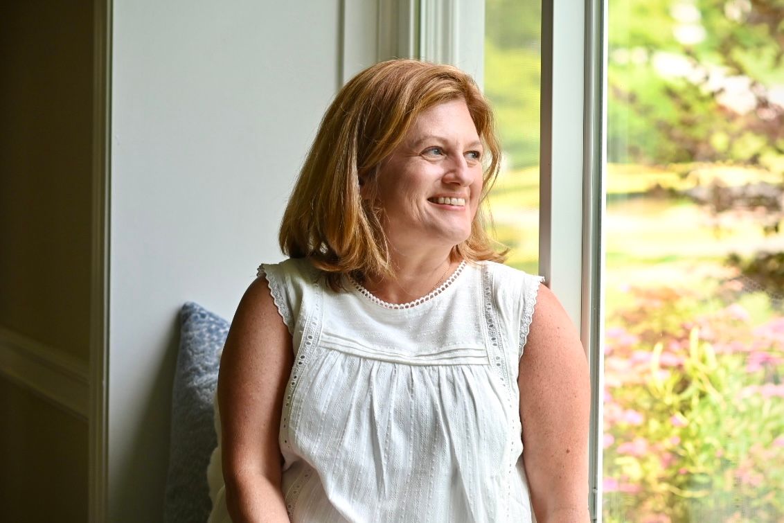 A smiling middle-aged woman with red hair stands by a sunlit window in a white sleeveless top, gazing outside.