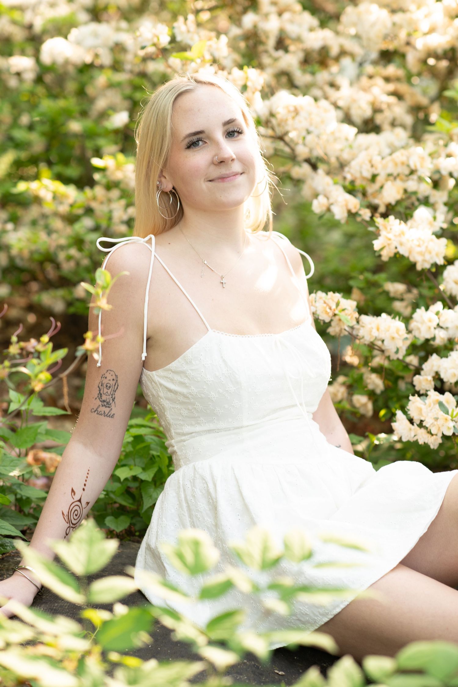 Blonde woman in white sundress sitting among blooming white flowers, showing arm tattoos and wearing a cross necklace.