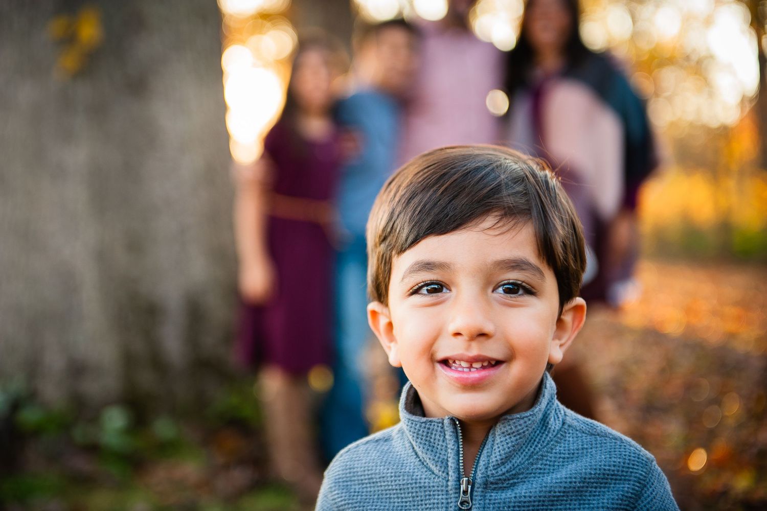 Smiling young boy in gray jacket poses outdoors during autumn family photo session with bokeh background.