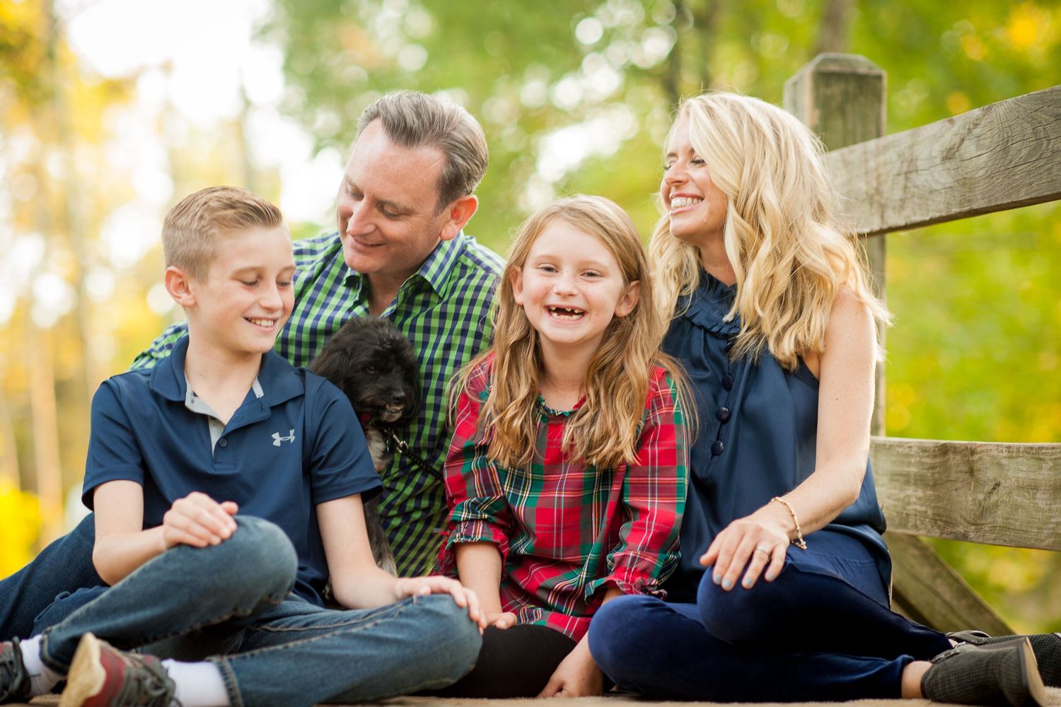 Happy family of four with a black puppy sitting on a wooden fence outdoors during fall season.