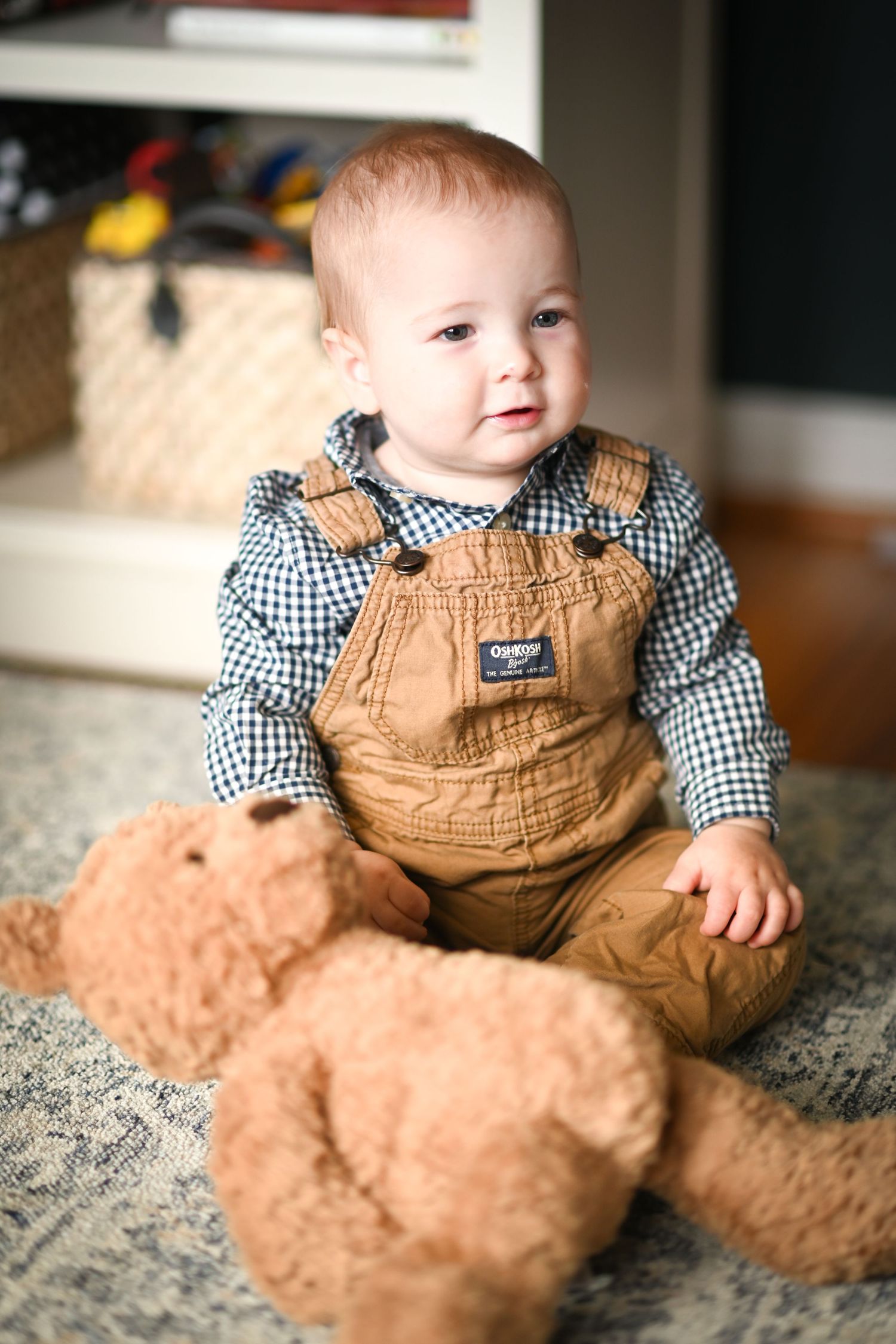 Adorable baby boy in OshKosh overalls and plaid shirt sitting with a large teddy bear on a rug.