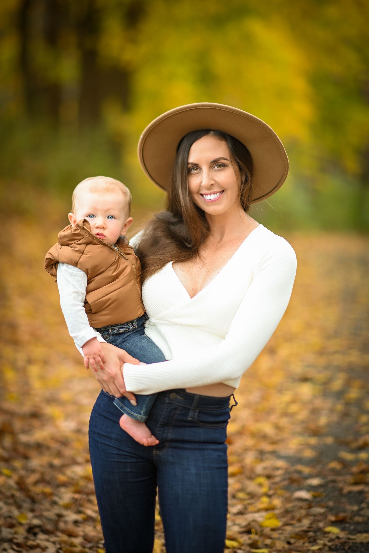 A smiling woman in a white top, jeans, and tan hat holds a baby in a brown jacket outdoors in an autumn park.
