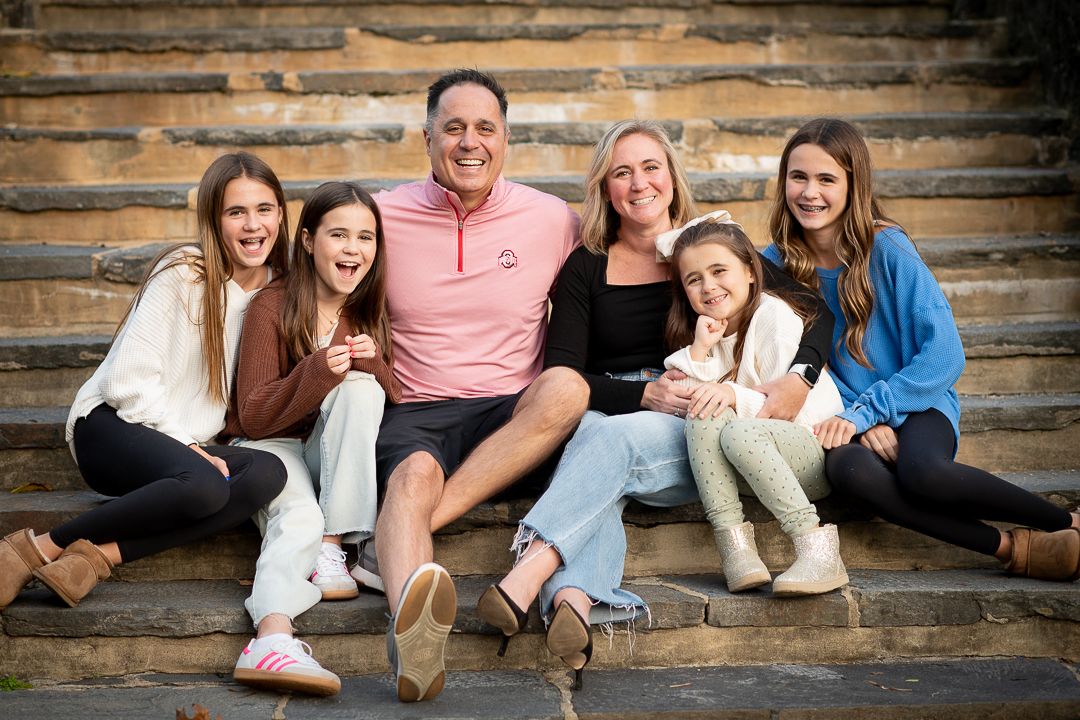 Happy family of six smiling together on stone steps outdoors, featuring parents and four daughters in casual attire.