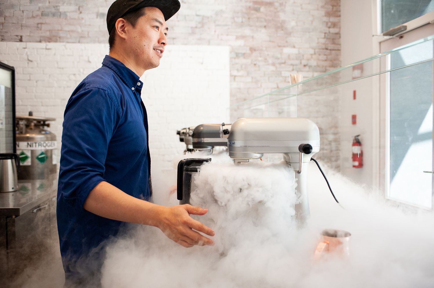 man in a blue shirt stands kitchen mixer as smoke pours out onto the counter