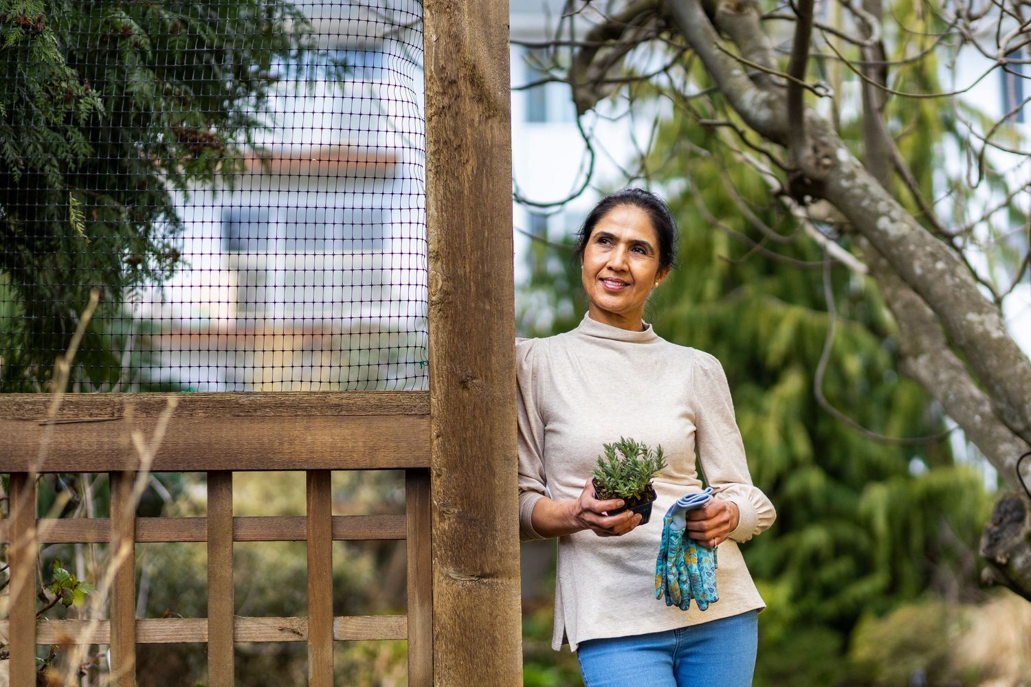 South Asian woman leaning on a wooden fence holding a pair of gardening gloves
