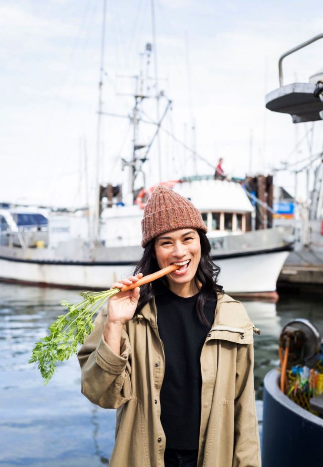 Young woman in a knitted hat eating a carrot with a boat in the background