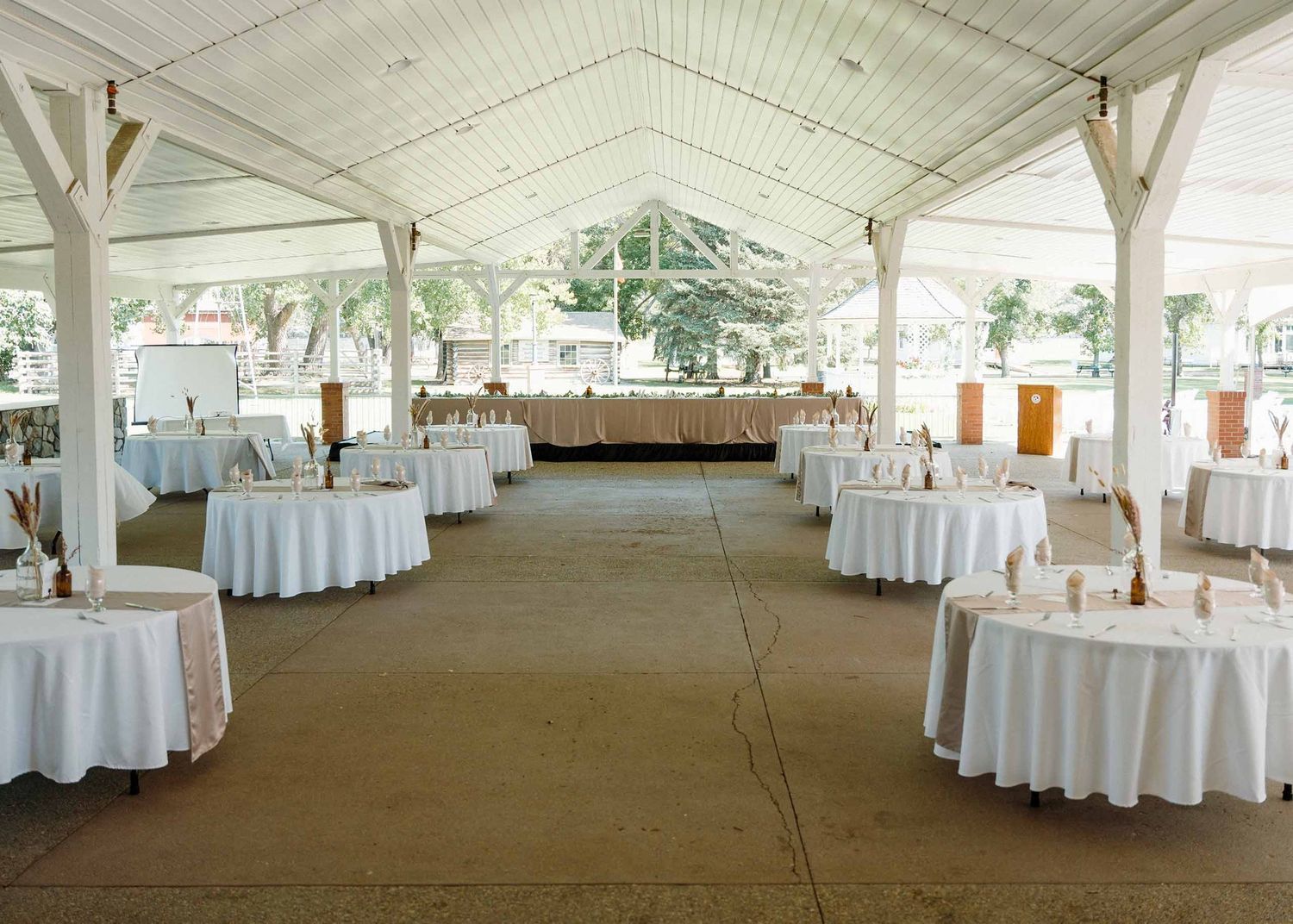 Elegant outdoor wedding reception venue with white round tables under a large open-air pavilion with white wooden beams.