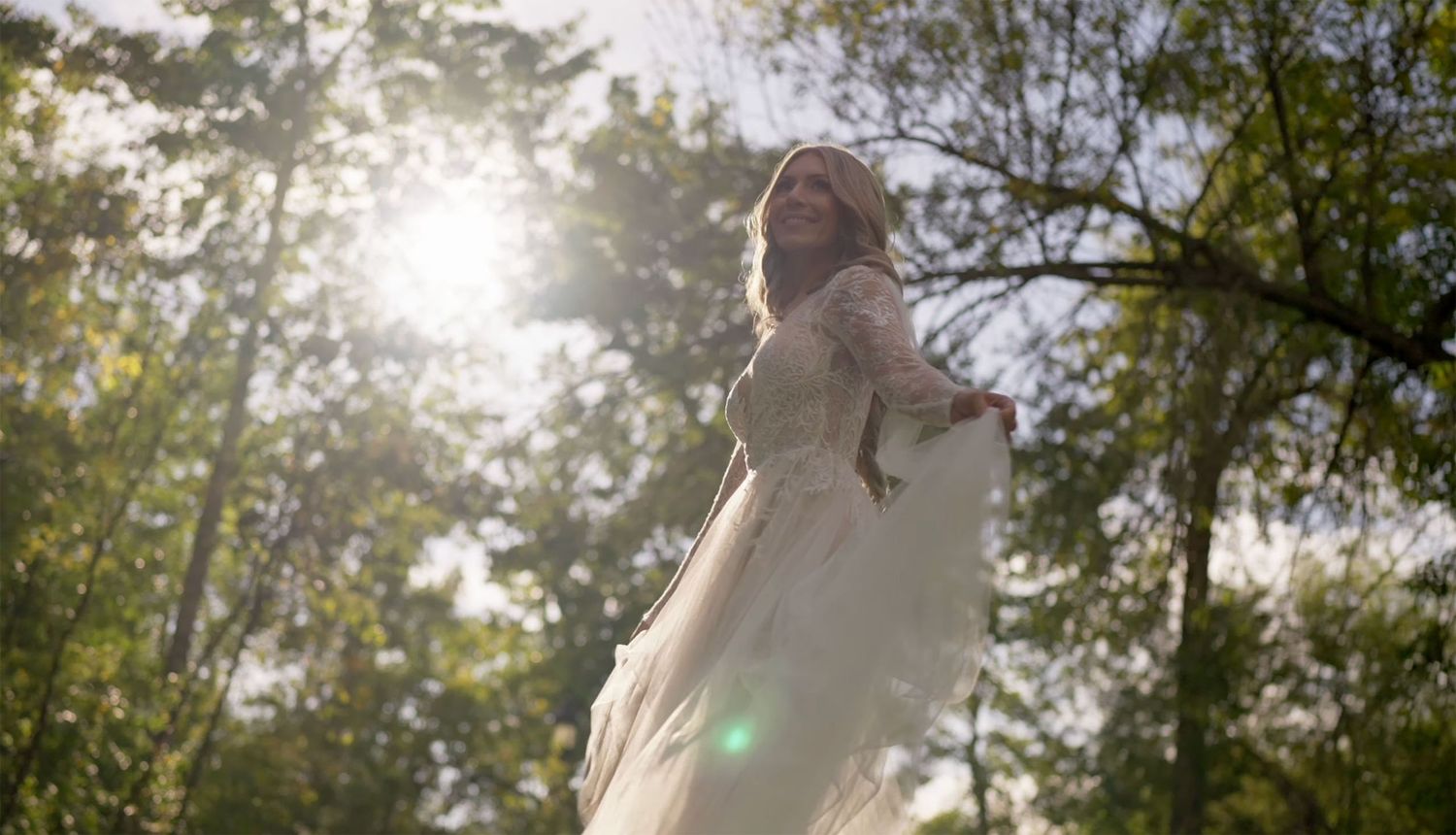 Woman in flowing white gown standing in sunlit forest, backlit by golden sun rays filtering through green trees.