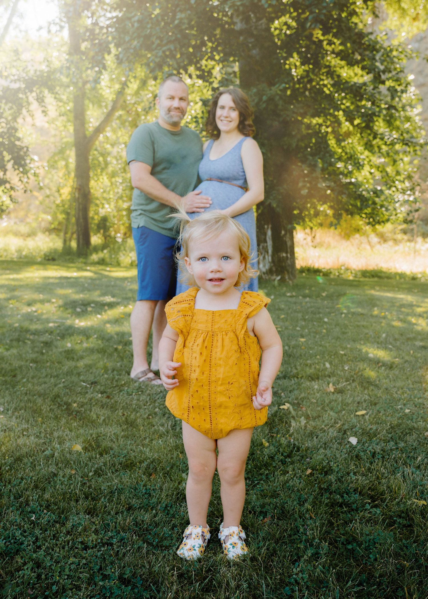 Smiling toddler in yellow romper stands in foreground as expecting parents embrace in golden sunlit park behind her.