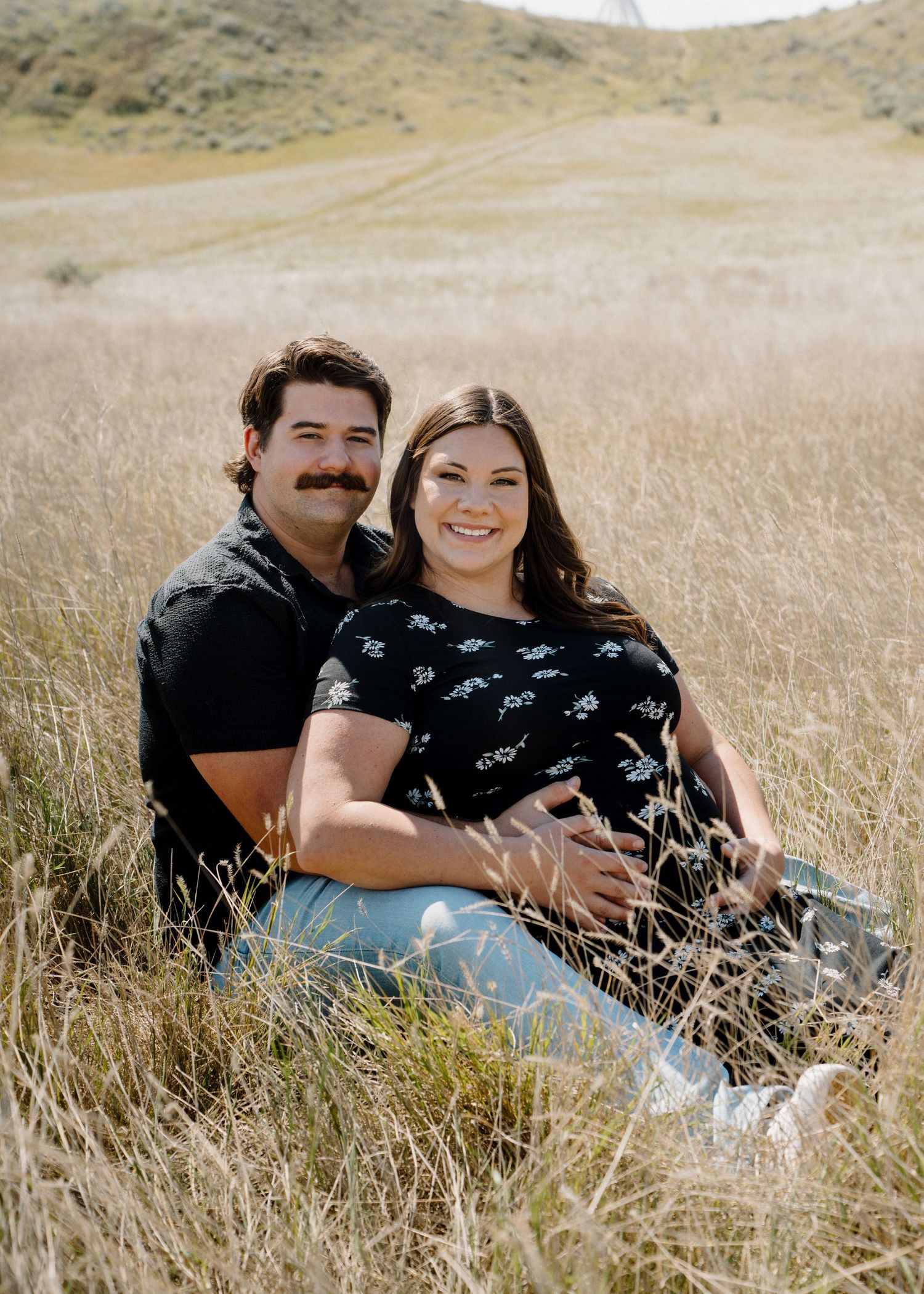 Expecting couple sits together in a golden field of tall grass, smiling warmly at the camera.