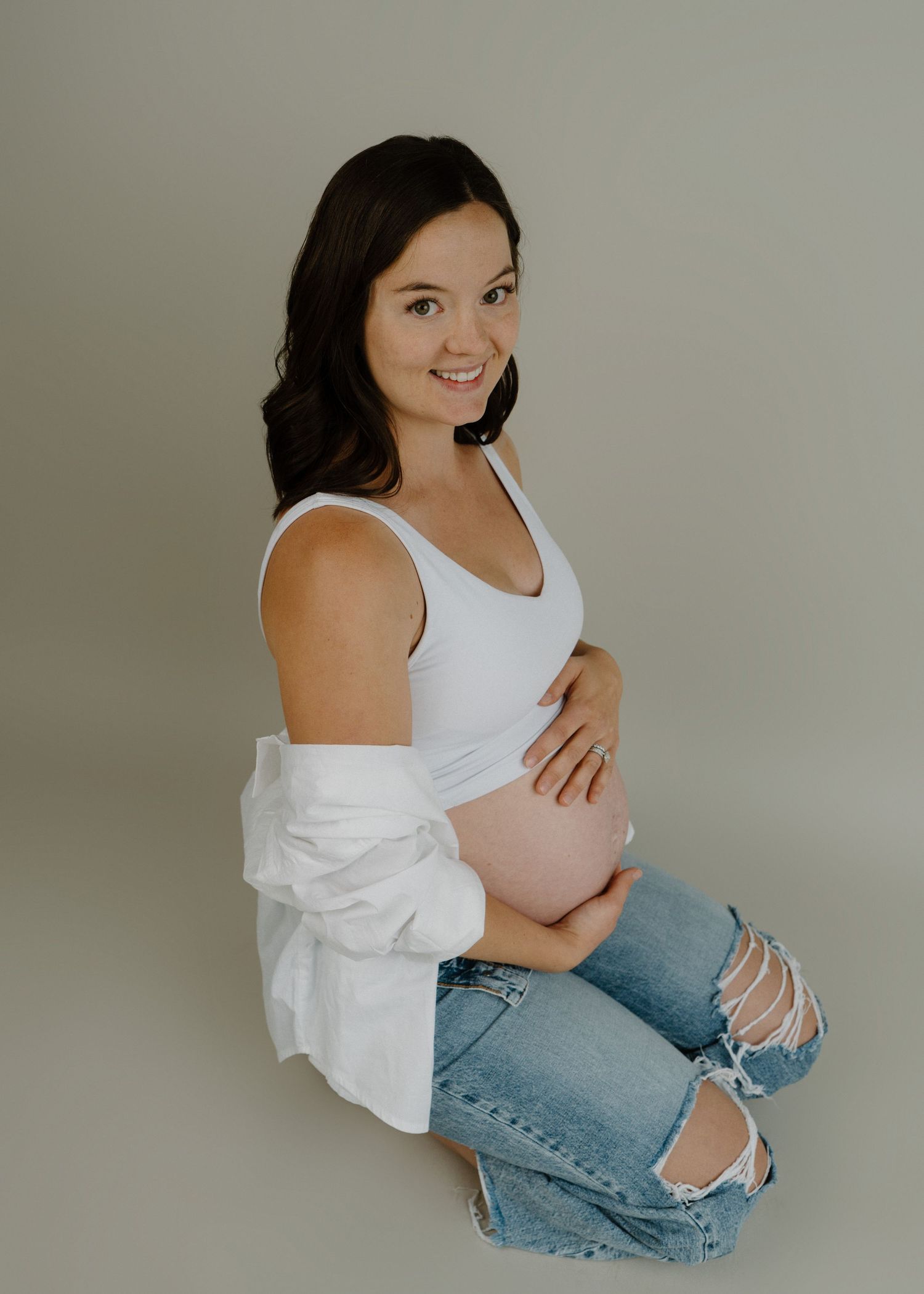 Pregnant woman in white tank and ripped jeans sits sideways cradling her baby bump in studio.