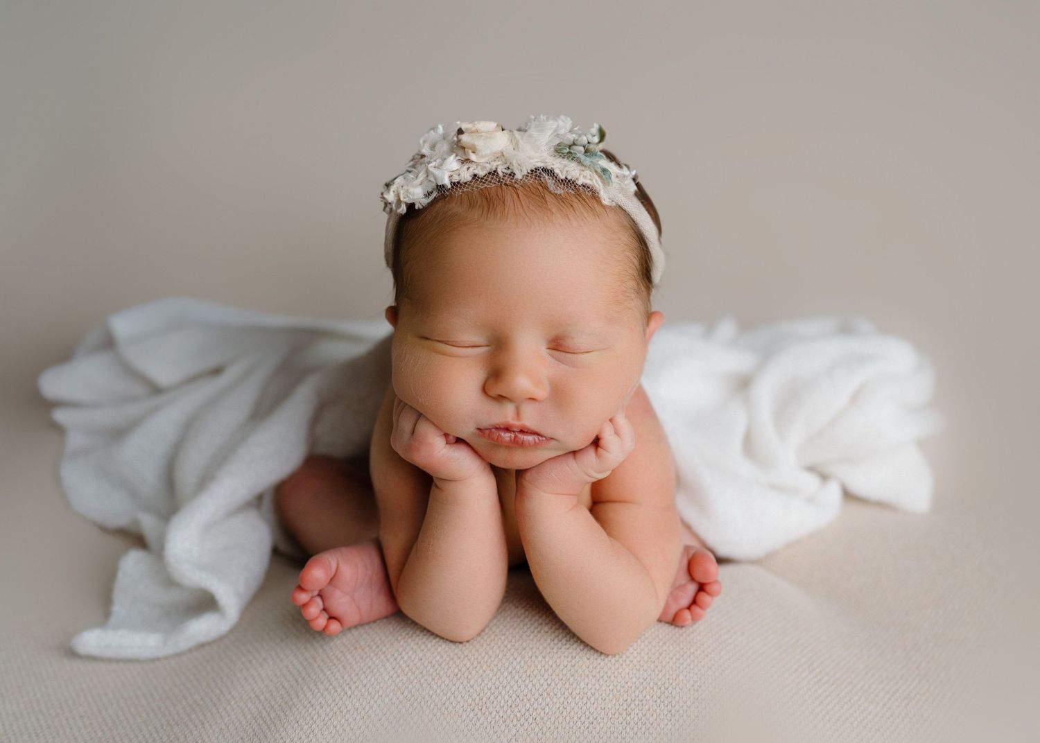 Peaceful sleeping newborn posed with hands under chin on white wrap wearing a floral crown headband.