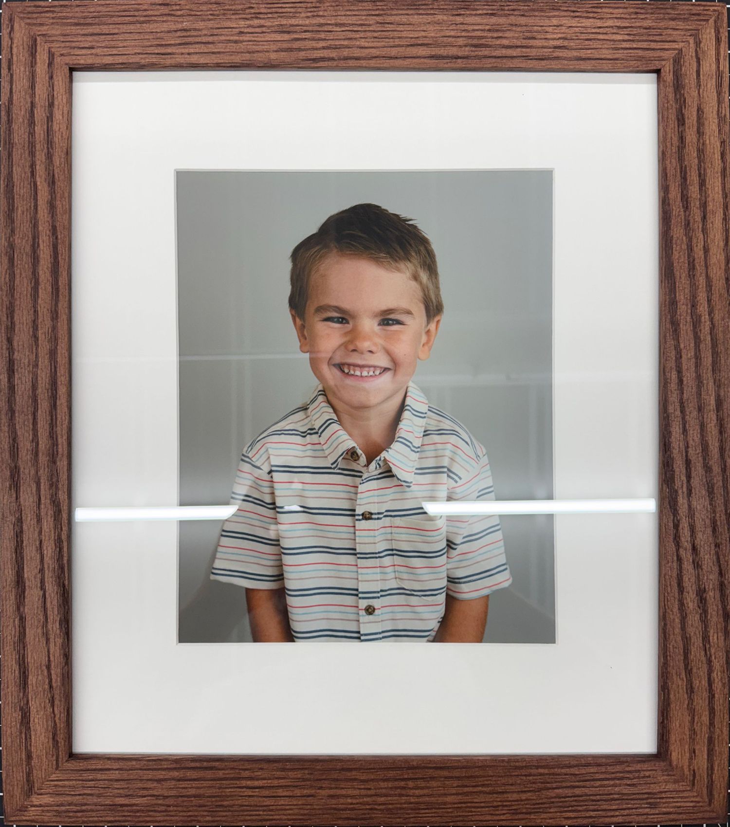 Framed school portrait of a smiling young boy wearing a striped polo shirt, displayed in a brown wooden frame.