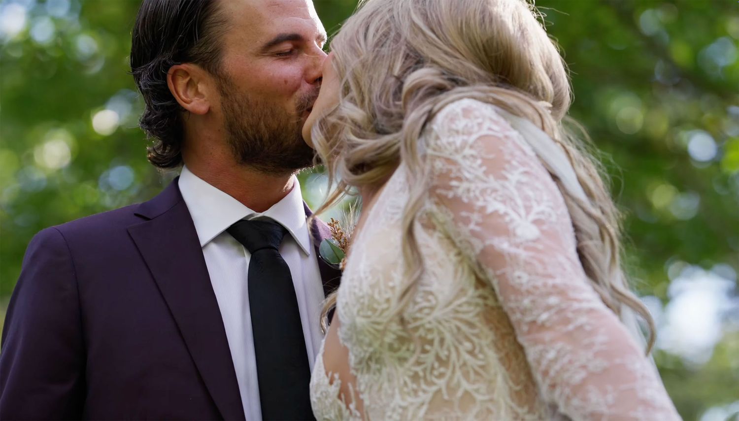 Couple sharing a romantic kiss at their wedding, groom in burgundy suit, bride in lace dress, outdoors.