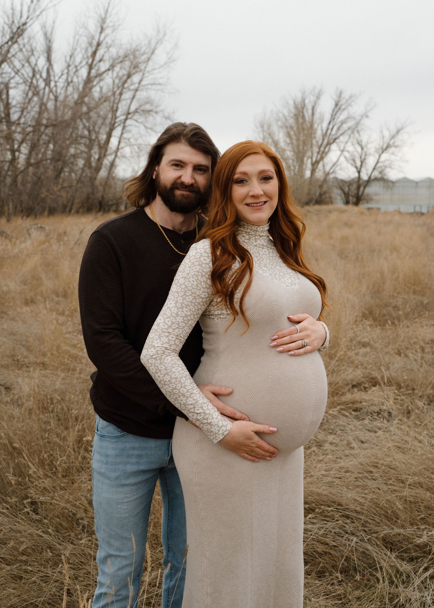 Bearded man embracing pregnant redheaded woman in beige lace dress in autumn field.