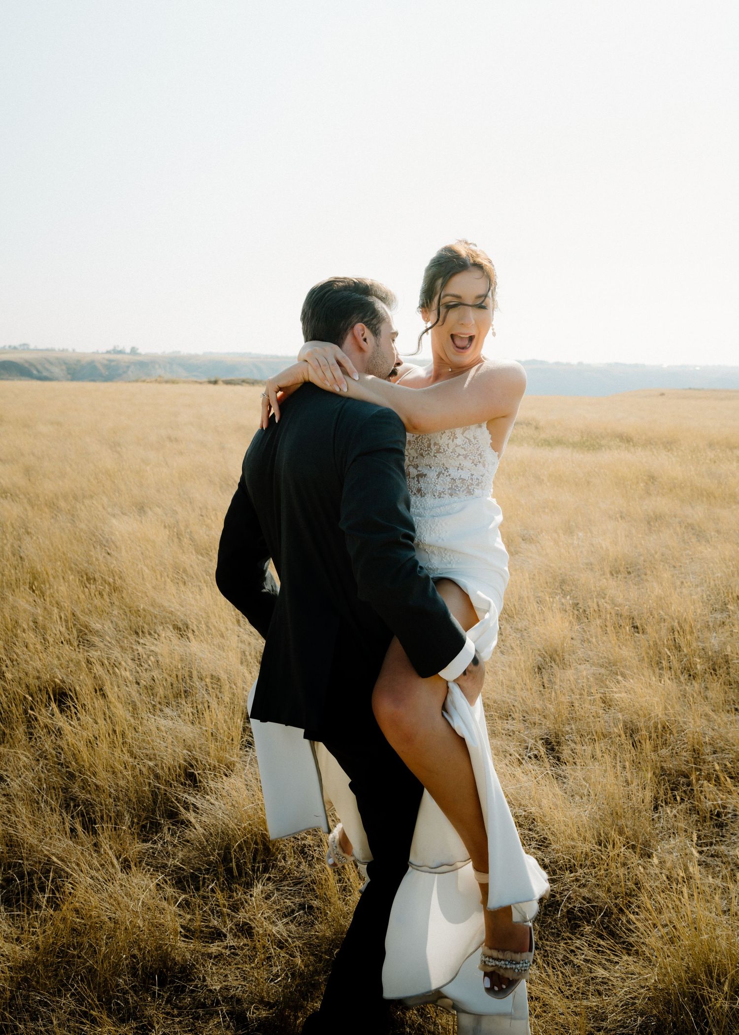 Groom lifts laughing bride in a golden wheat field during sunny outdoor wedding portraits.