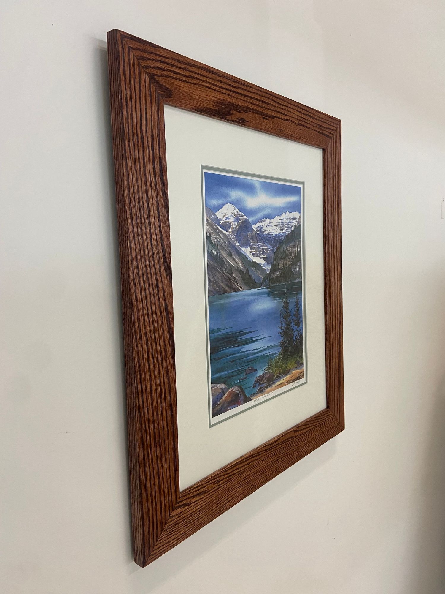 Framed landscape painting of a mountain lake with snow-capped peaks in a warm oak wood frame.