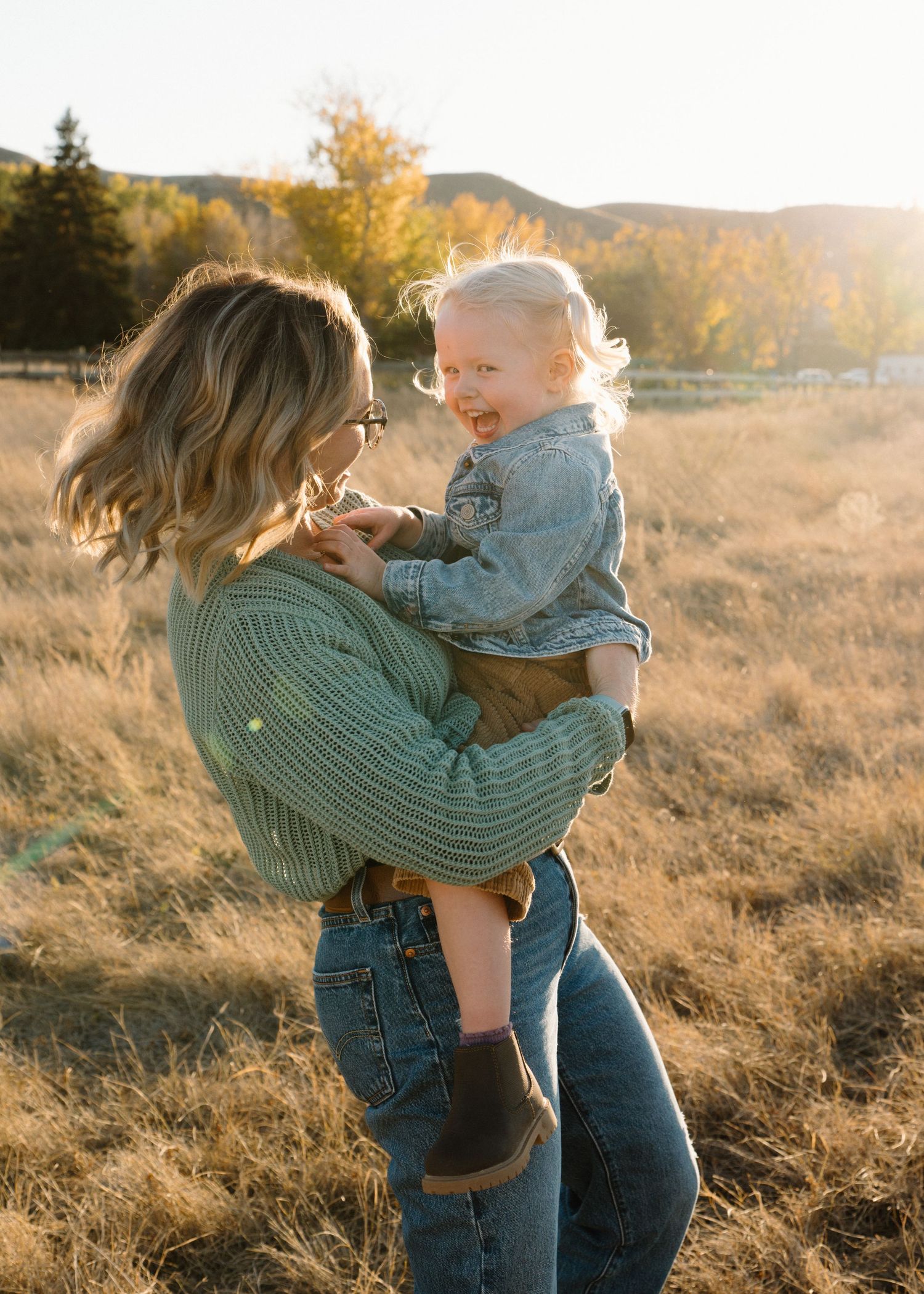 Joyful blonde toddler laughs while being lifted by mom in a sunlit golden meadow during fall.