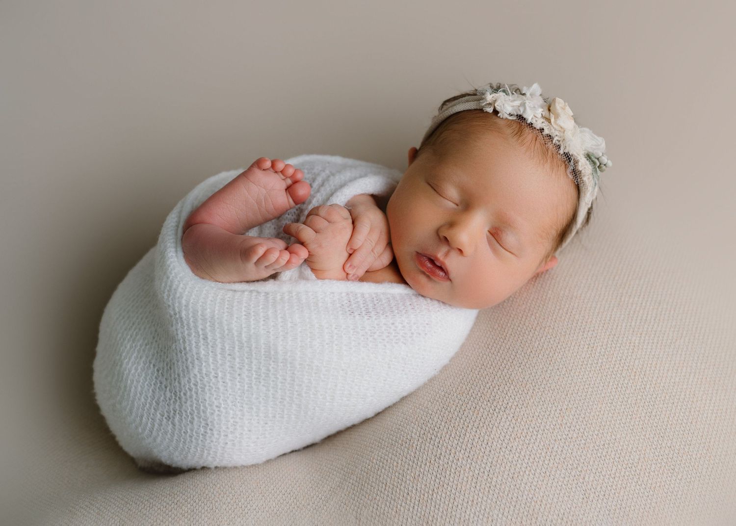 Sleeping newborn baby swaddled in white wrap with floral headband on beige backdrop.