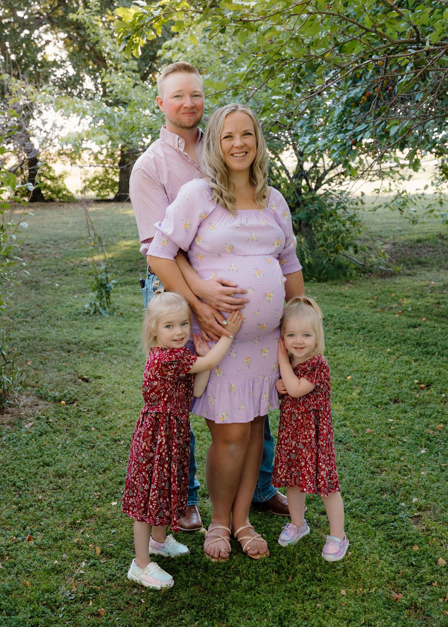 Family maternity portrait with parents and two girls in red dresses under green trees in garden.