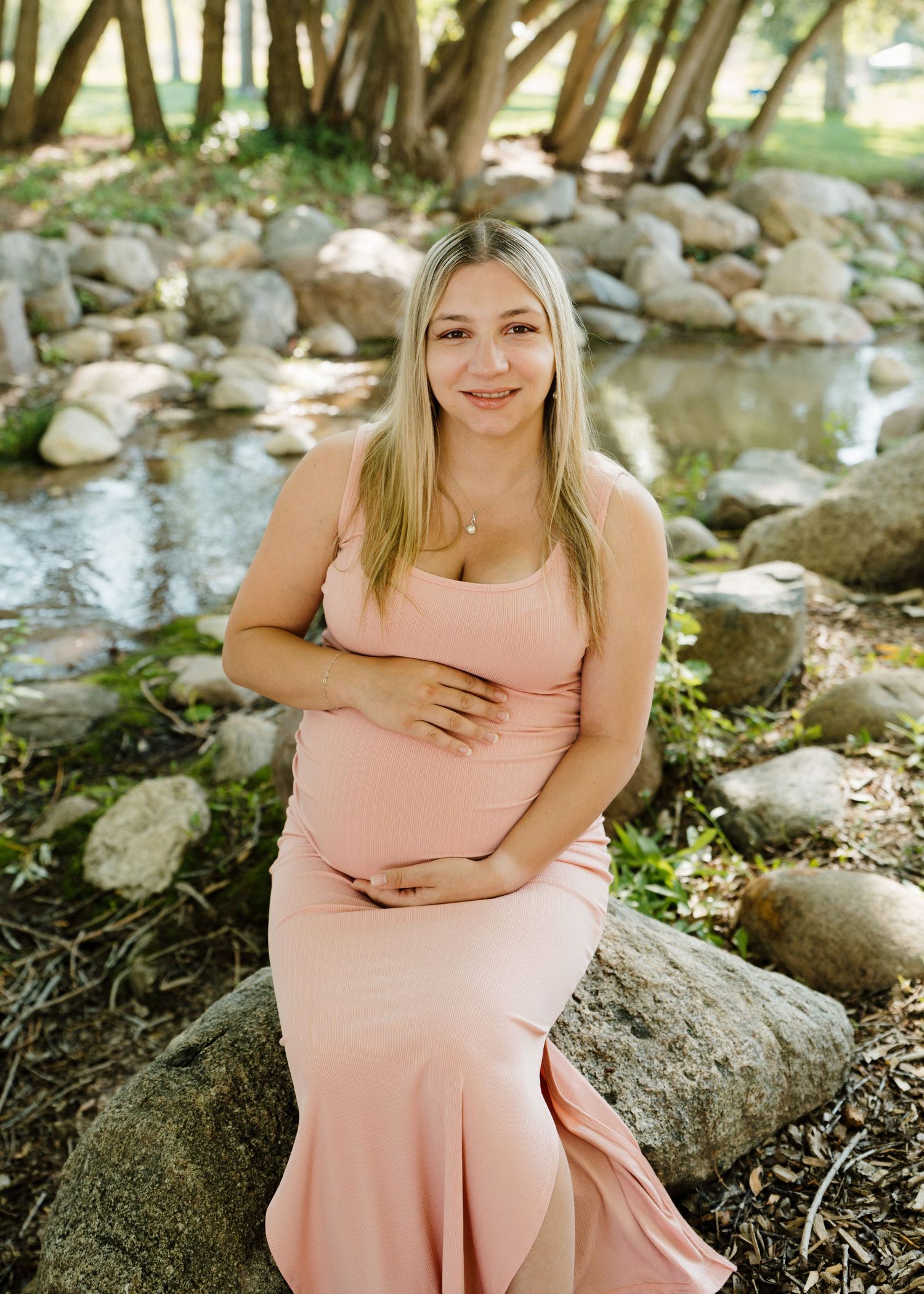 Pregnant woman in pink maternity gown sitting on rocks by a stream in a serene outdoor setting.