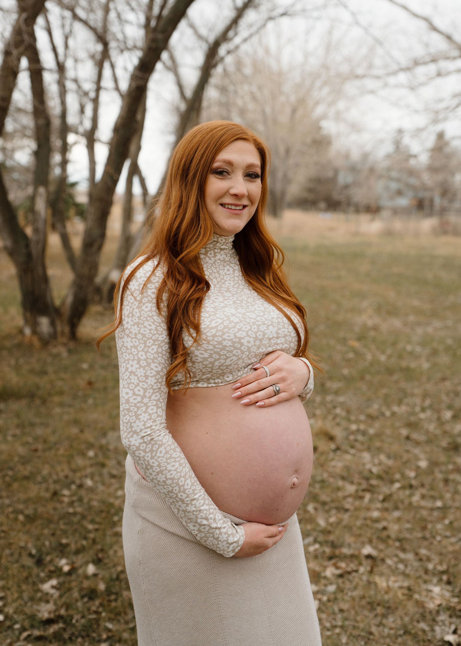Smiling pregnant redhead woman in floral crop top and beige skirt cradling baby bump outdoors.