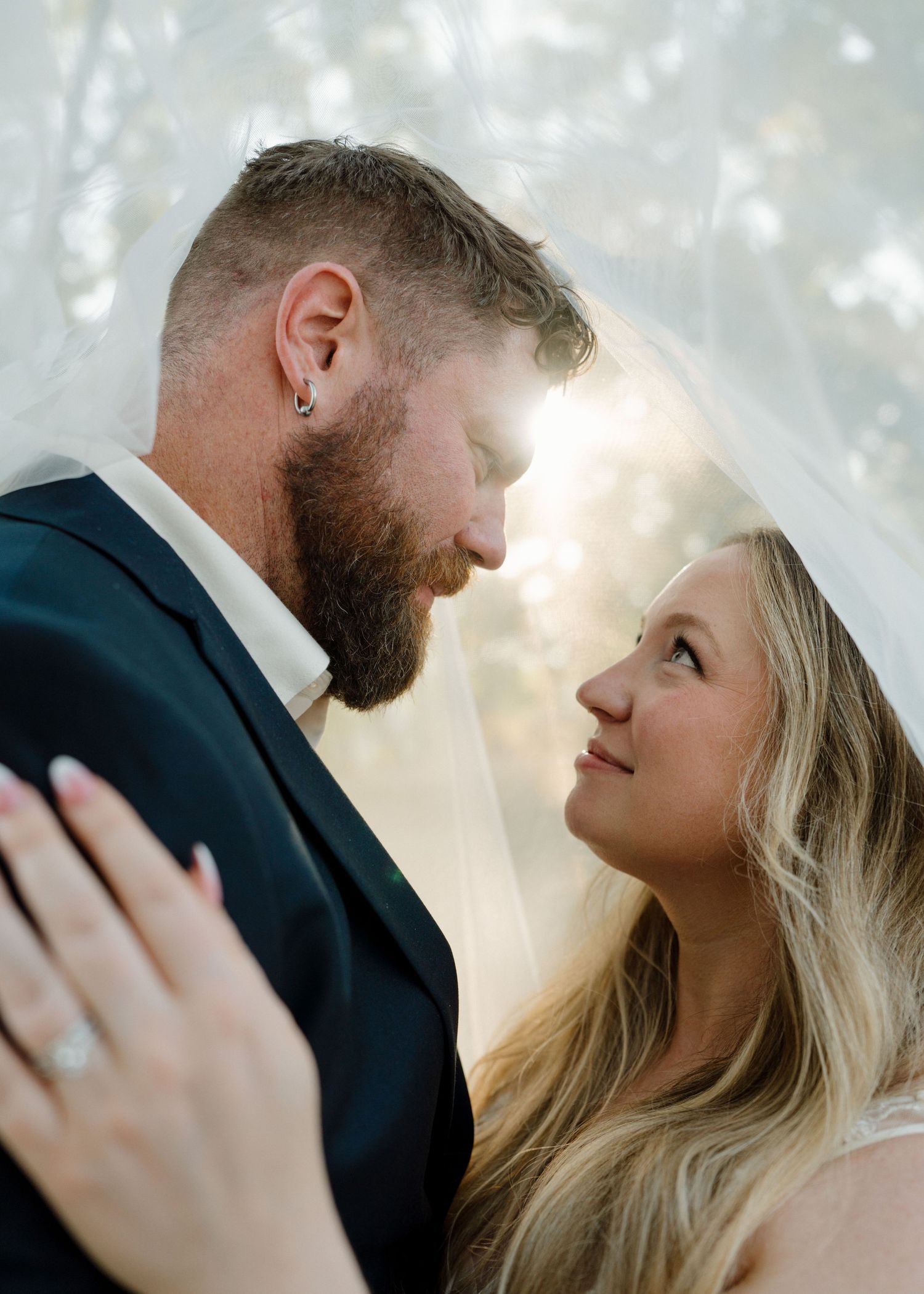 Bride and groom sharing an intimate moment under a veil with soft backlight in an outdoor setting.