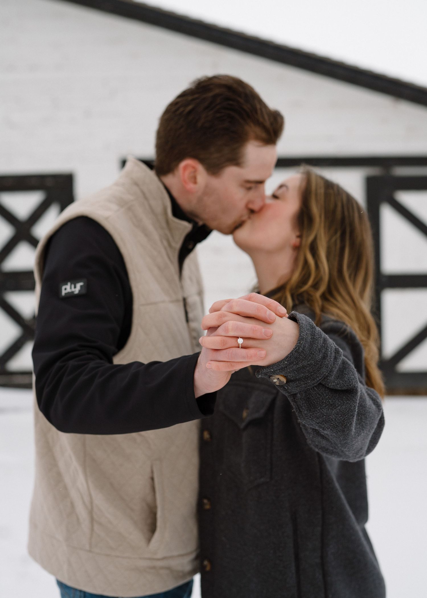 Couple sharing a romantic kiss in the snow outside a white barn, showing off engagement ring.
