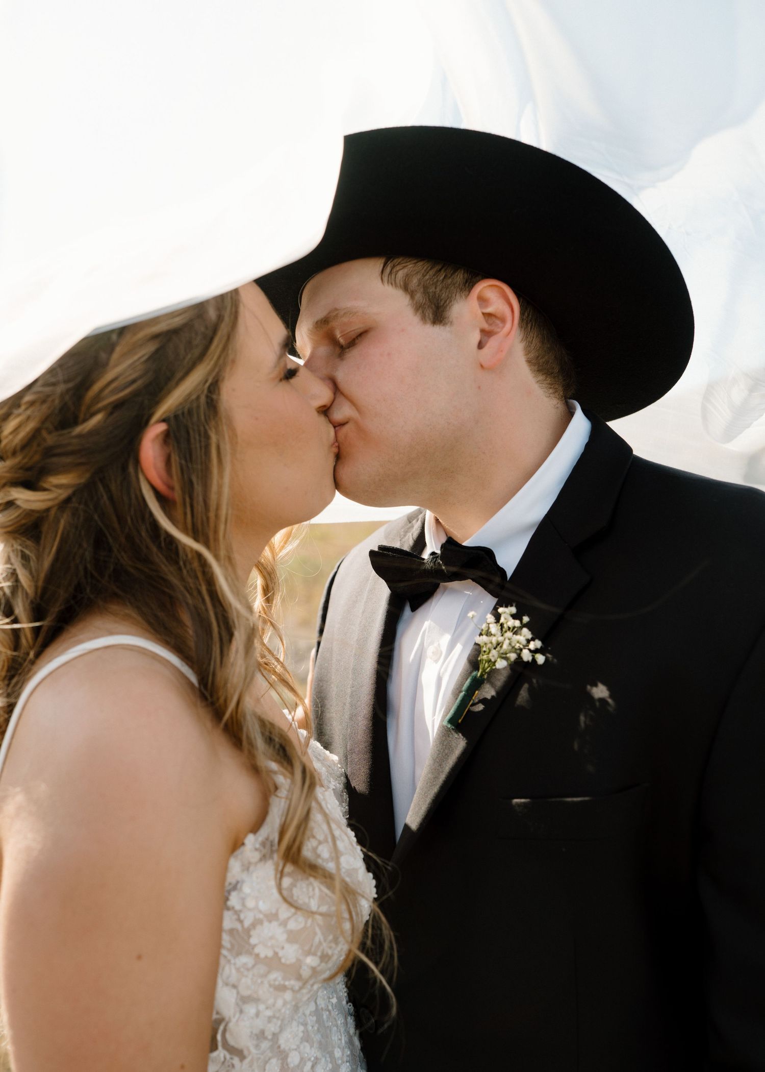 Bride and groom sharing romantic kiss, groom in black cowboy hat and tuxedo with boutonniere.