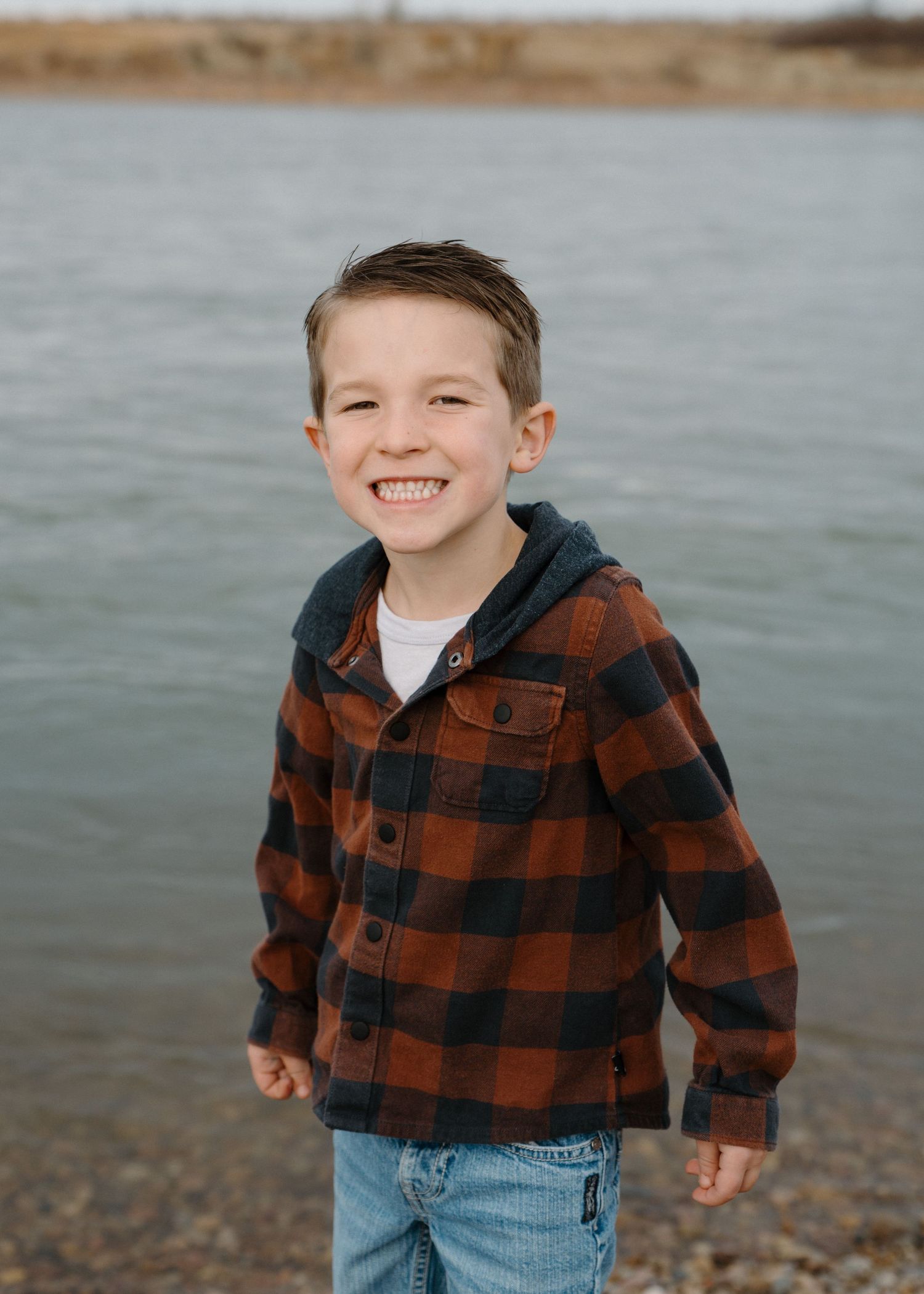 Smiling young boy in orange plaid flannel jacket standing near a lake on a cloudy day.