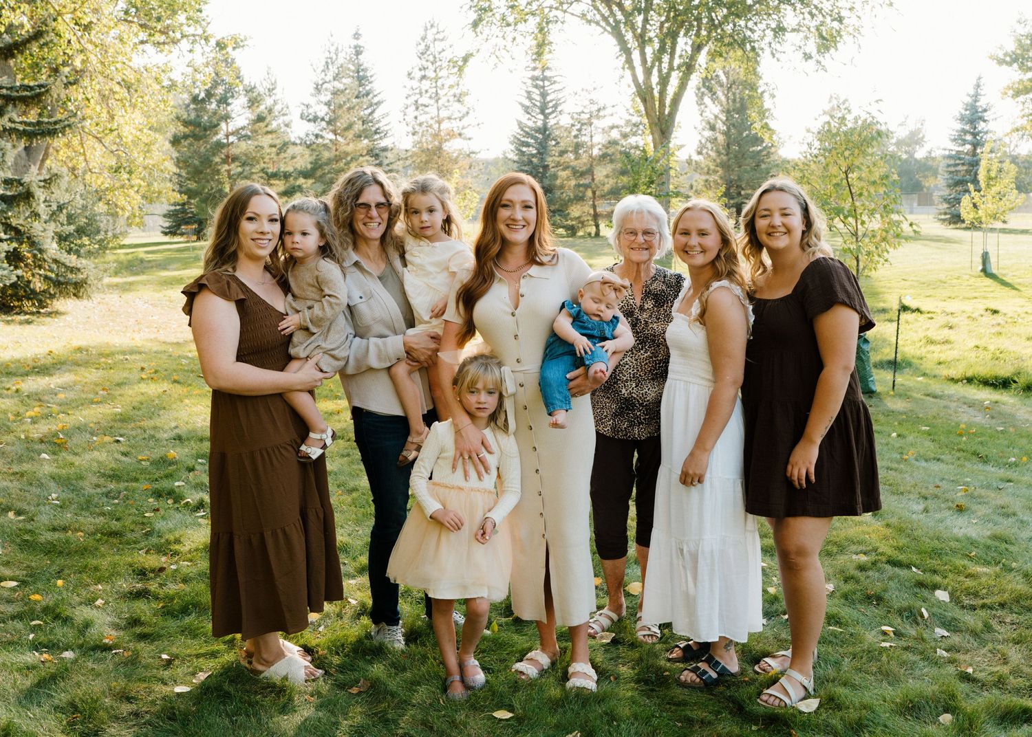 Group of women and children smiling outdoors on green grass with trees in background.