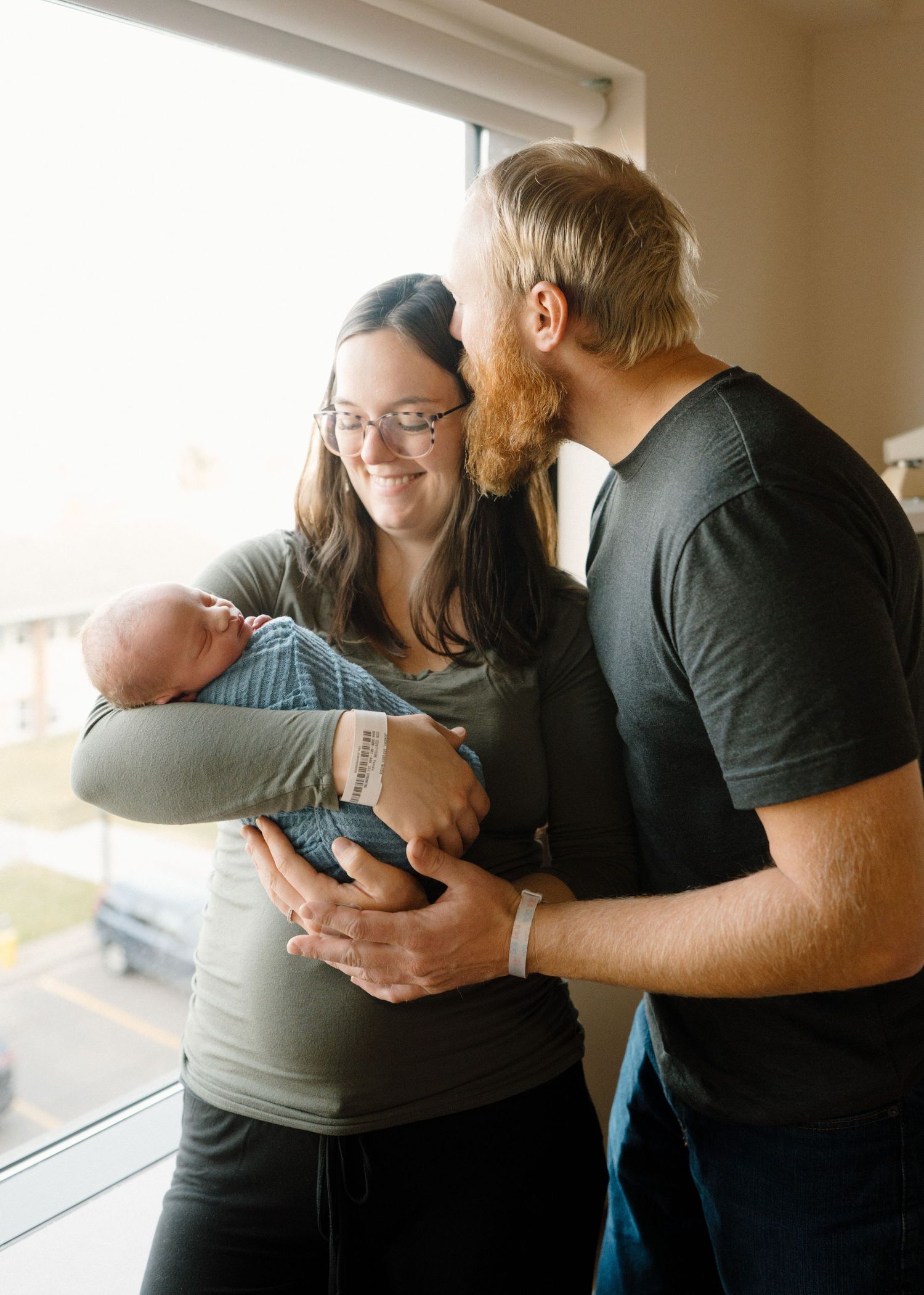 Happy new parents smiling and bonding with their newborn baby by hospital room window after delivery.