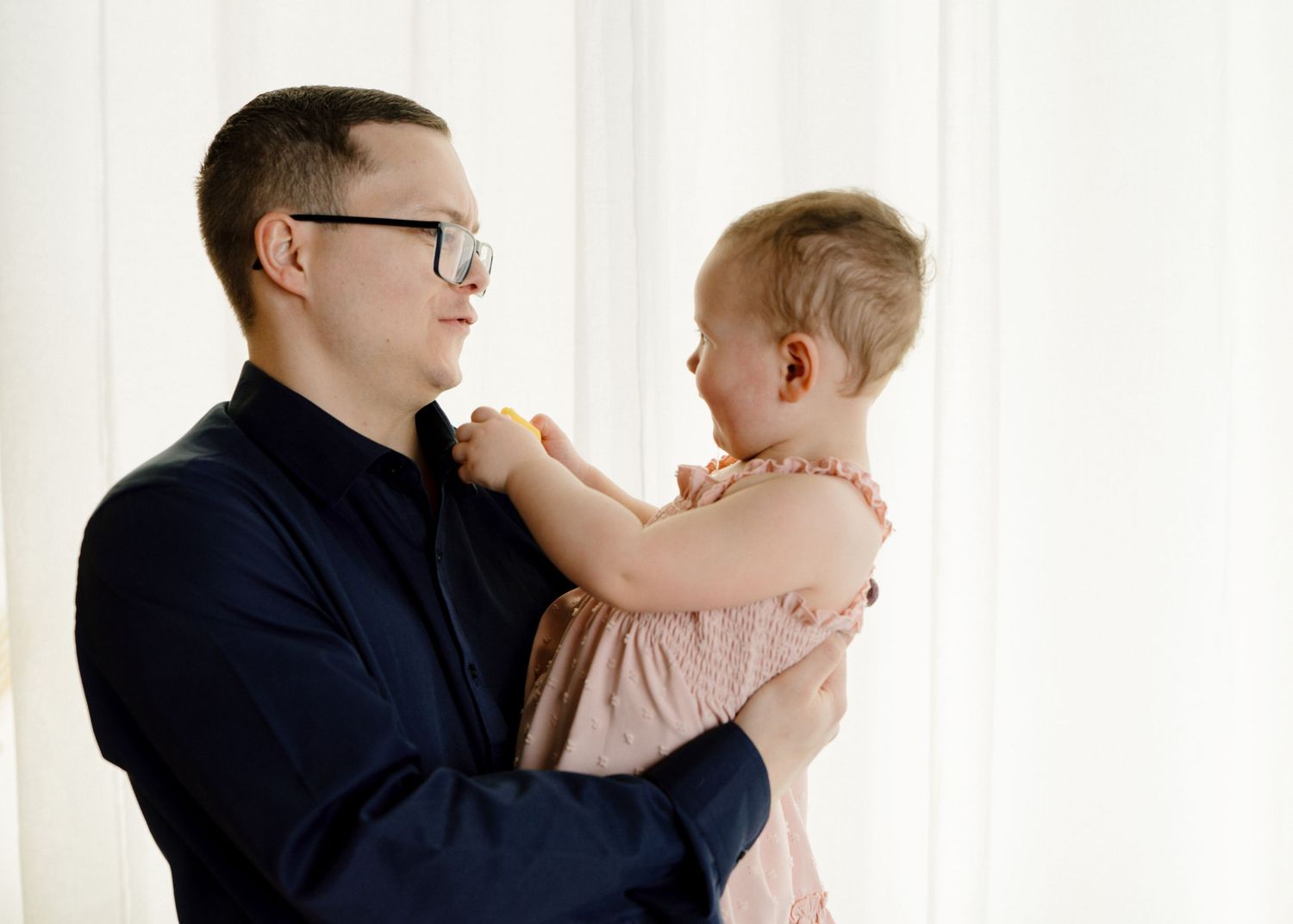 Father wearing glasses holds and smiles at his baby girl in front of white curtains.