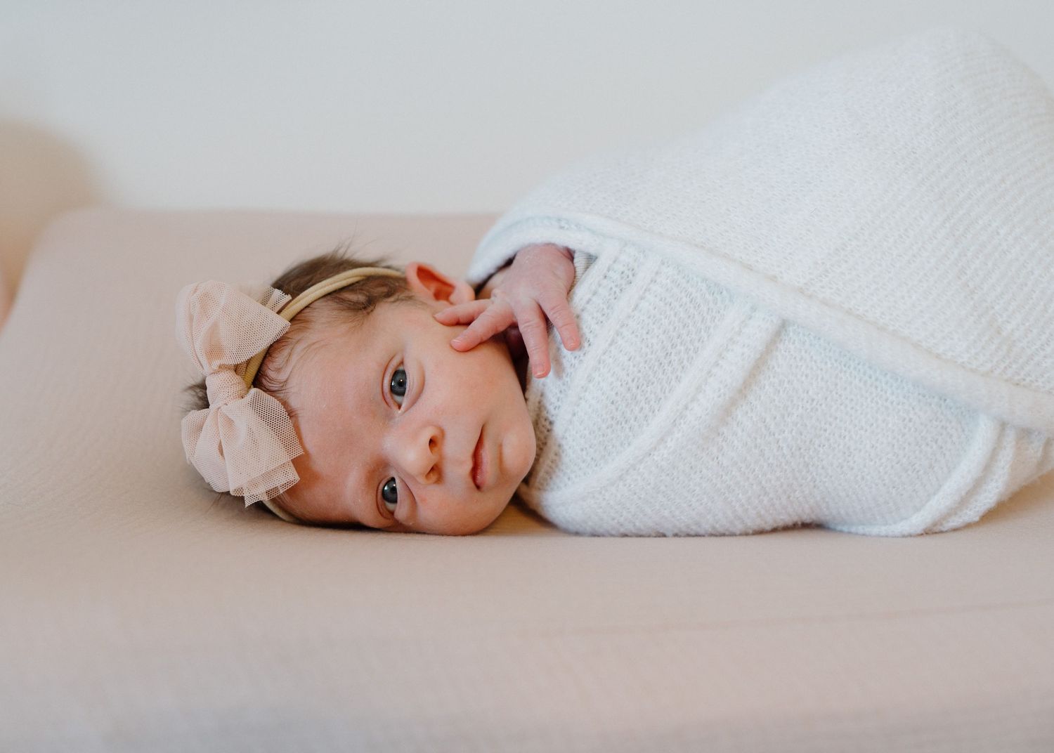 Newborn baby girl swaddled in white wrap with pink bow headband lying on beige surface.