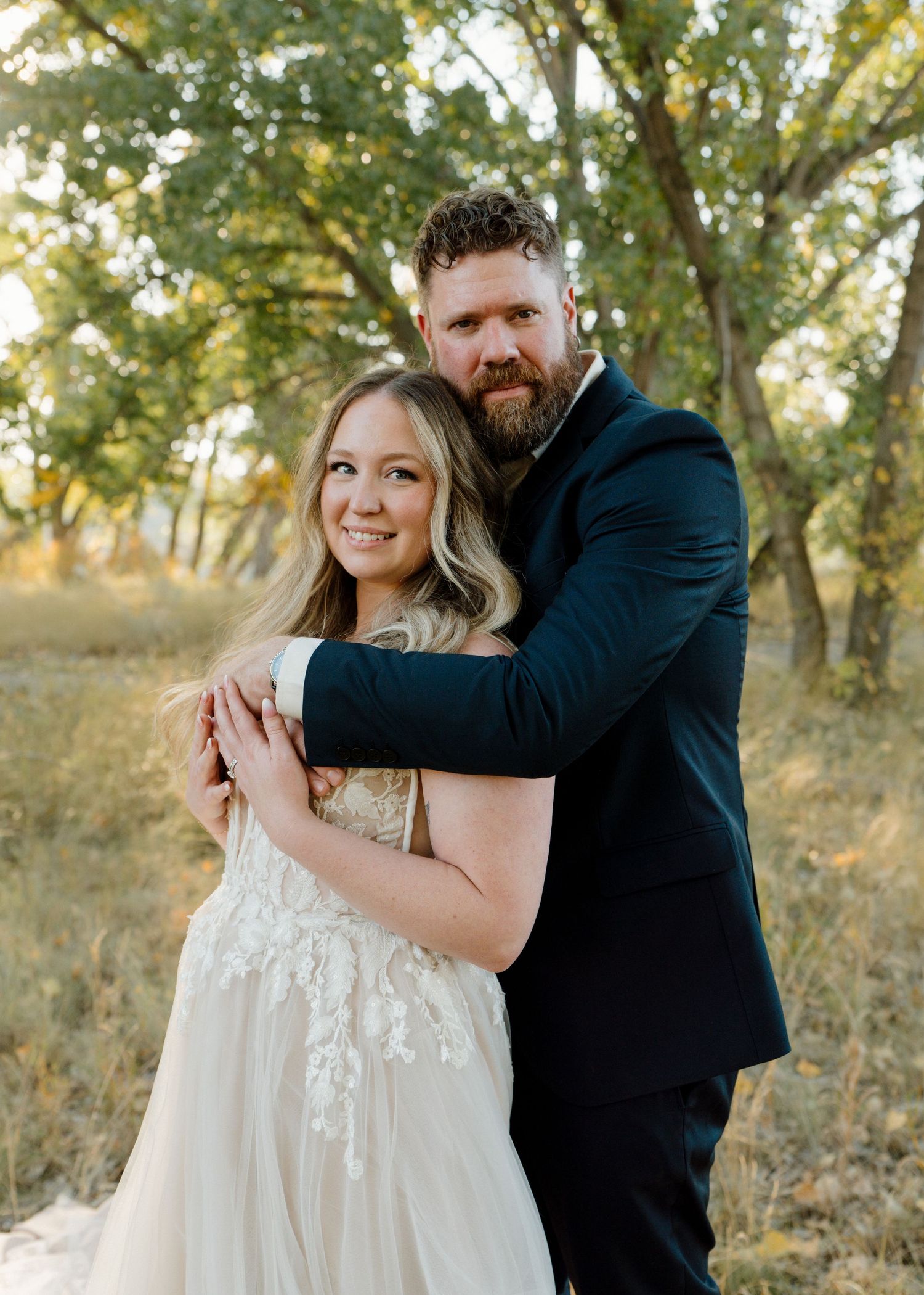 Bride in lace gown and bearded groom in navy suit embracing in a sunlit wooded outdoor setting.