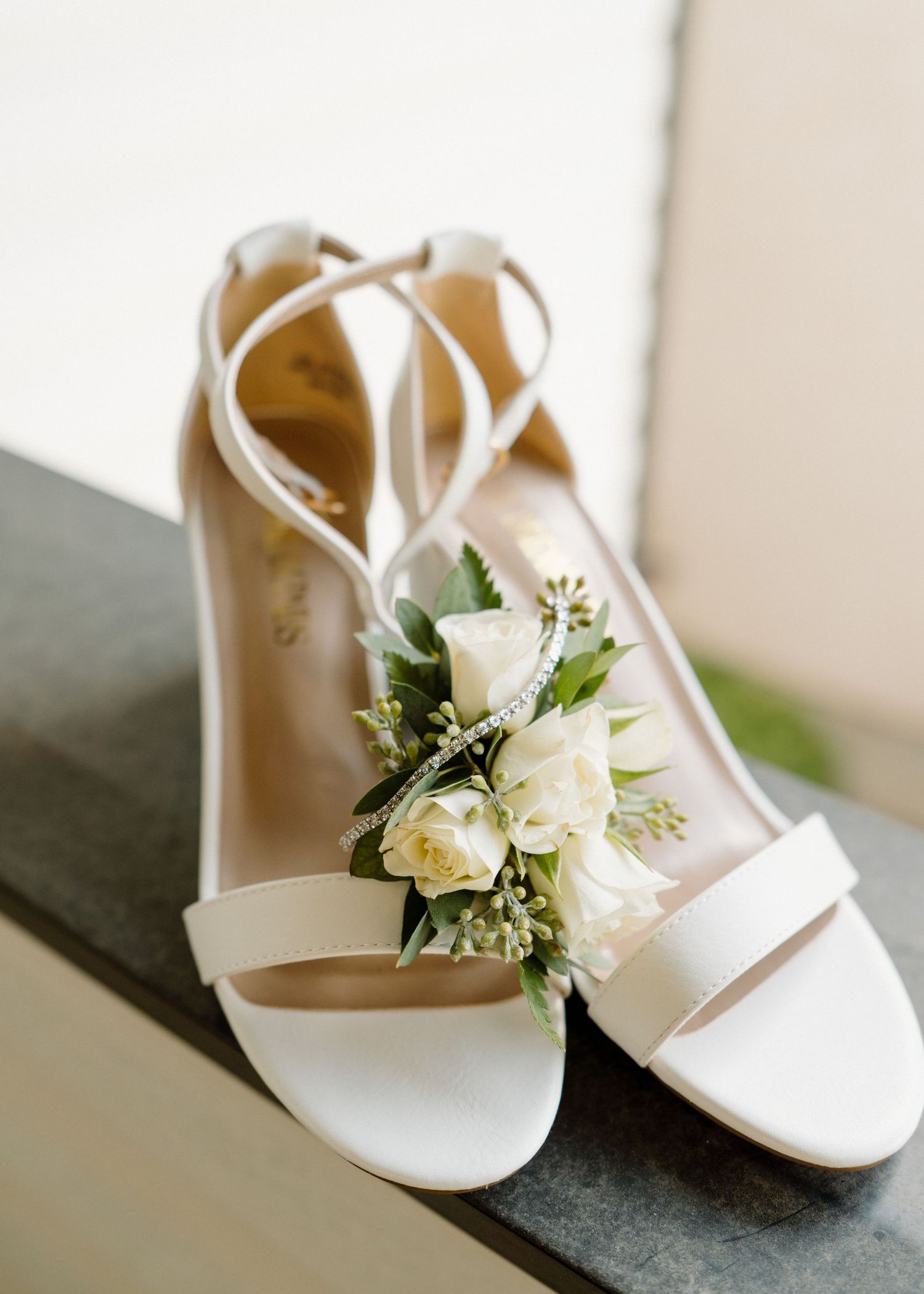 White bridal heels with floral corsage featuring roses and greenery displayed on stone surface.