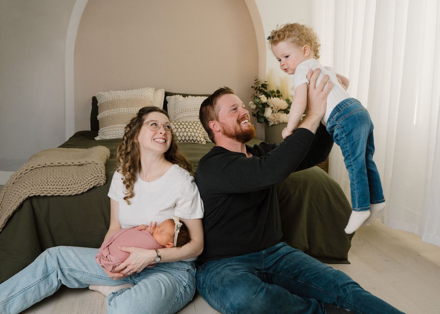 Happy family sitting on floor with dad lifting toddler while mom holds newborn baby during in-home session.