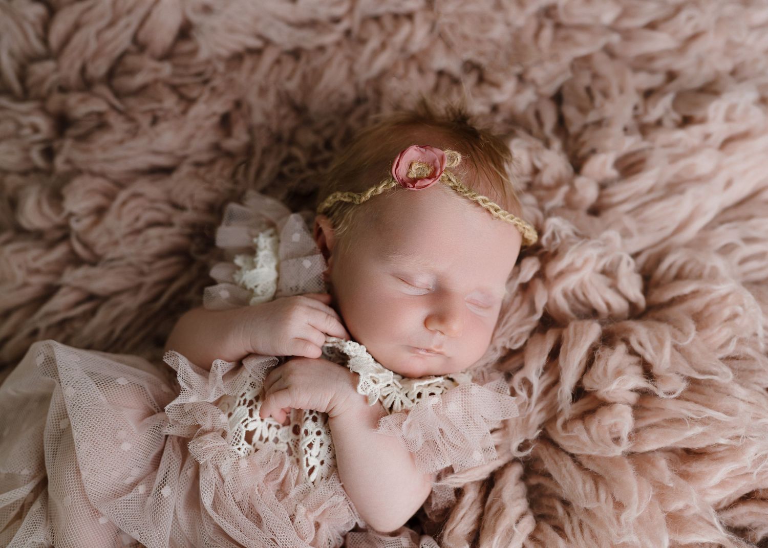 Newborn baby girl sleeping in pink lace outfit with rose headband on fluffy pink backdrop.